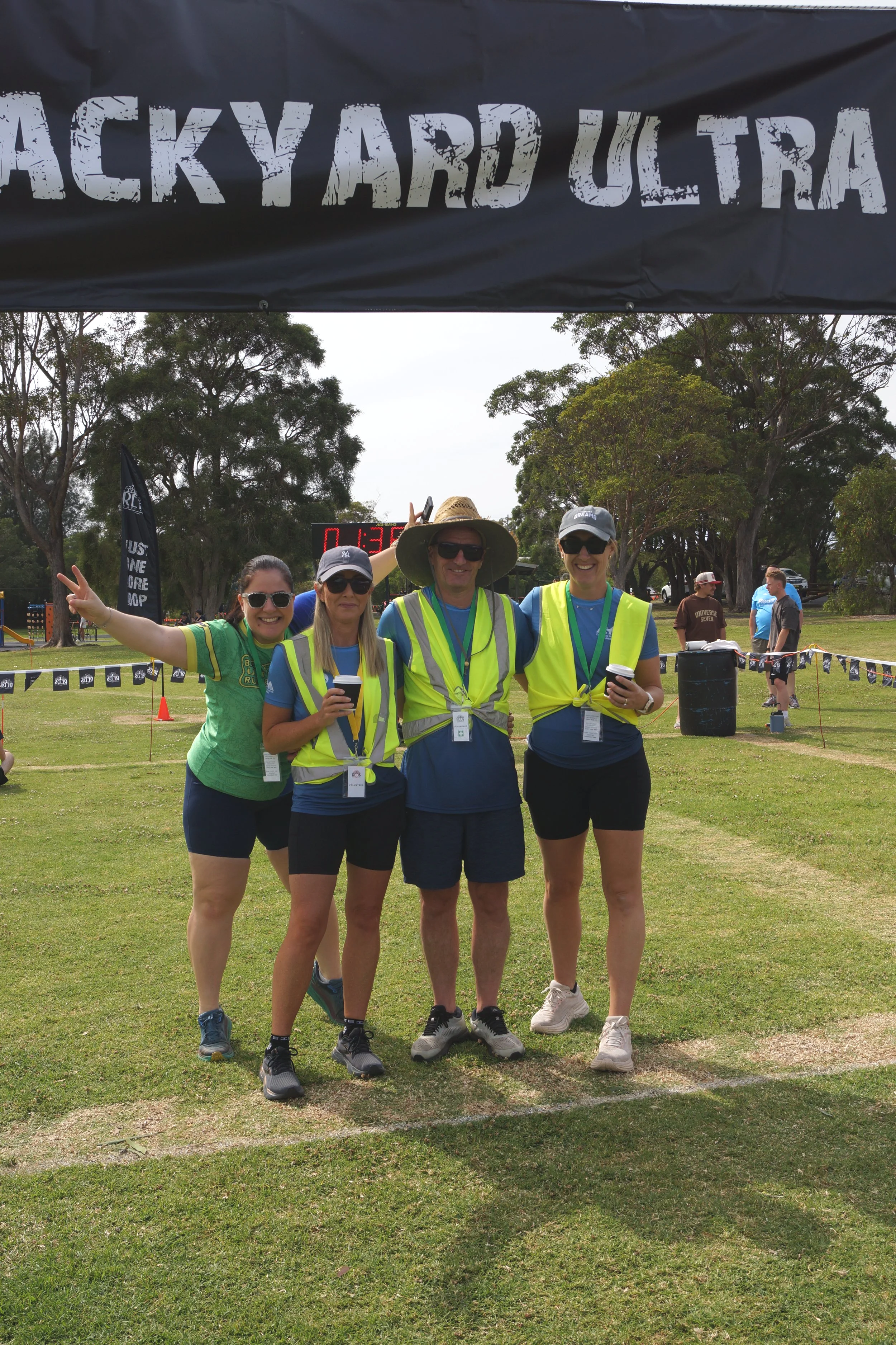 Four people standing under a banner that reads 'BACKYARD ULTRA,' smiling and posing for the photo during a race event in a park. They are wearing athletic clothing, sports watches, and high-visibility vests, with some holding cups. Two women are maki