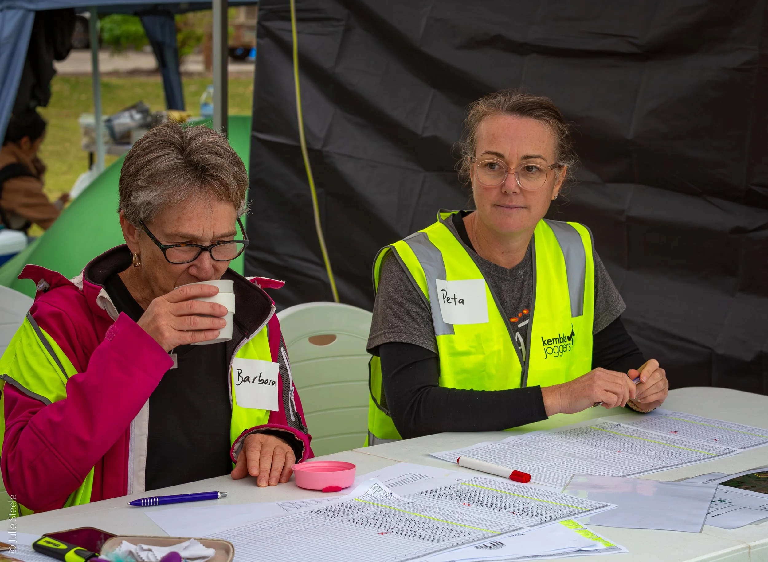 Two women sitting at a table with papers, pens, and other items, outdoors, one is drinking from a cup and wearing a pink jacket, the other is looking ahead with a yellow safety vest and a name tag that says 'Peta'.