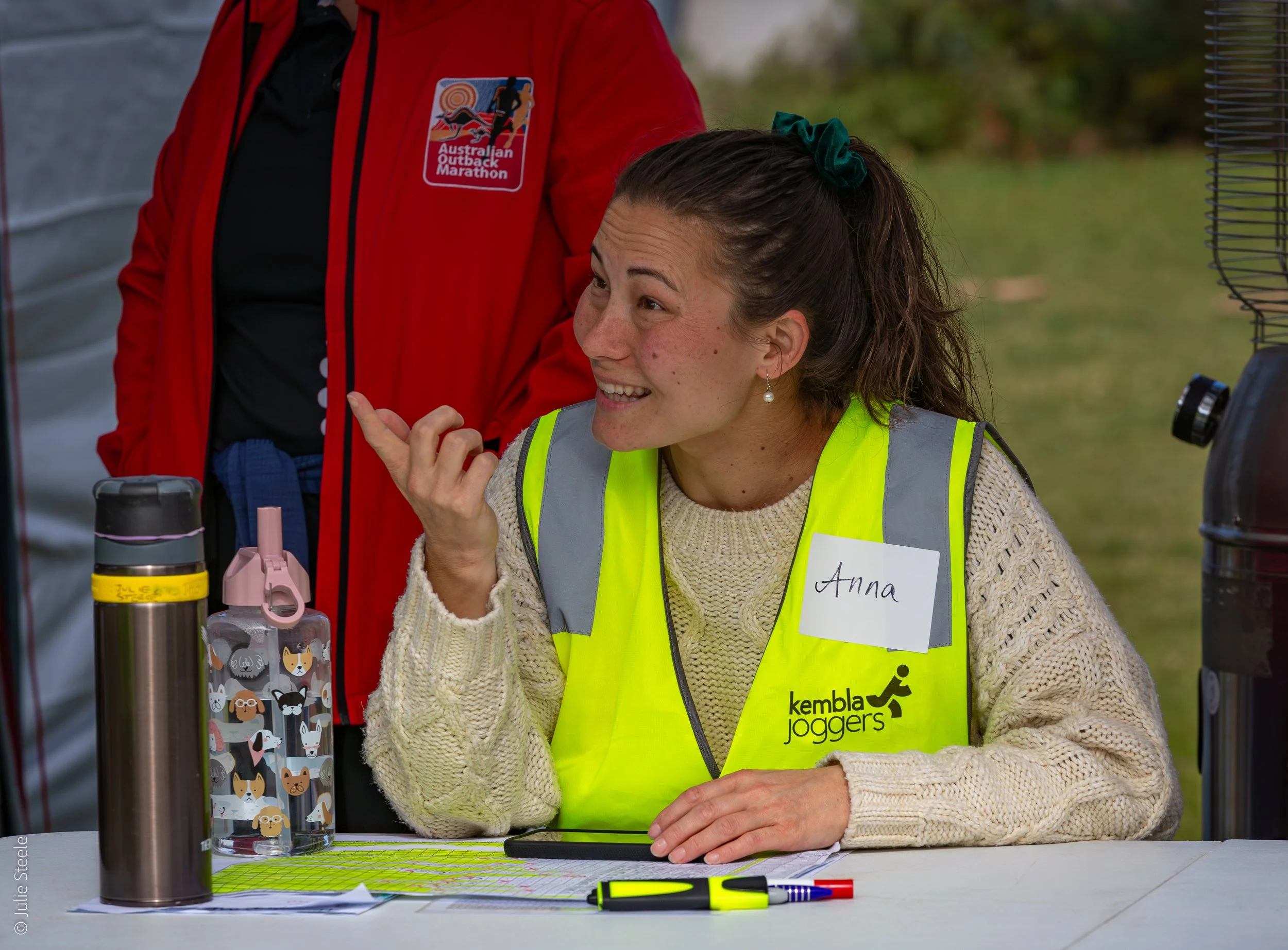 A woman named Anna wearing a yellow vest with 'kembla joggers' logo, sitting at a table with a water bottle, a pink water cup, and some papers, smiling and gesturing with her right hand. She has brown hair tied back with a green scrunchie, and is out