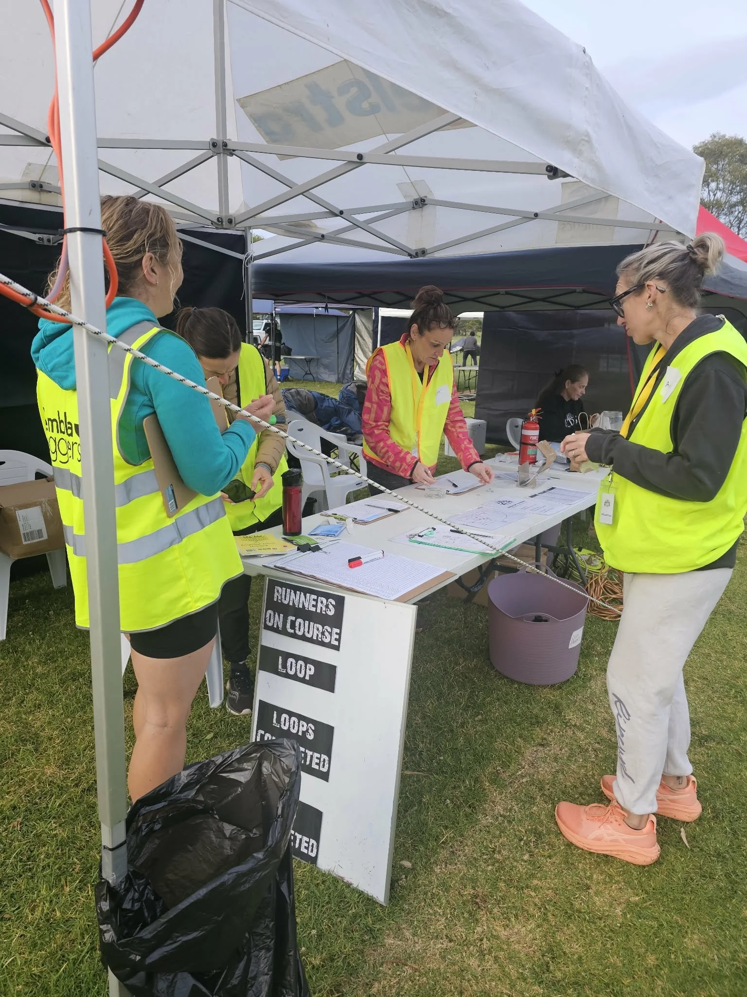 Race officials and volunteers in yellow vests preparing at a race registration table under a white canopy, with a sign indicating procedures for course runners and loops, and participants working nearby