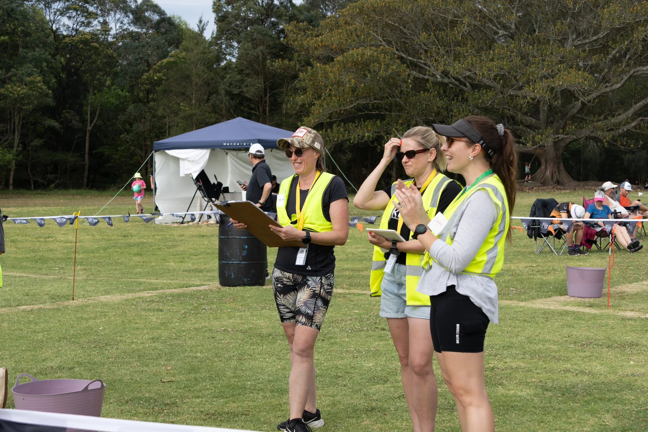 Three women wearing yellow safety vests at an outdoor event, standing on grass with trees in the background. One woman is holding a clipboard, another is adjusting her sunglasses, and the third is clapping. Several people are sitting and standing in 