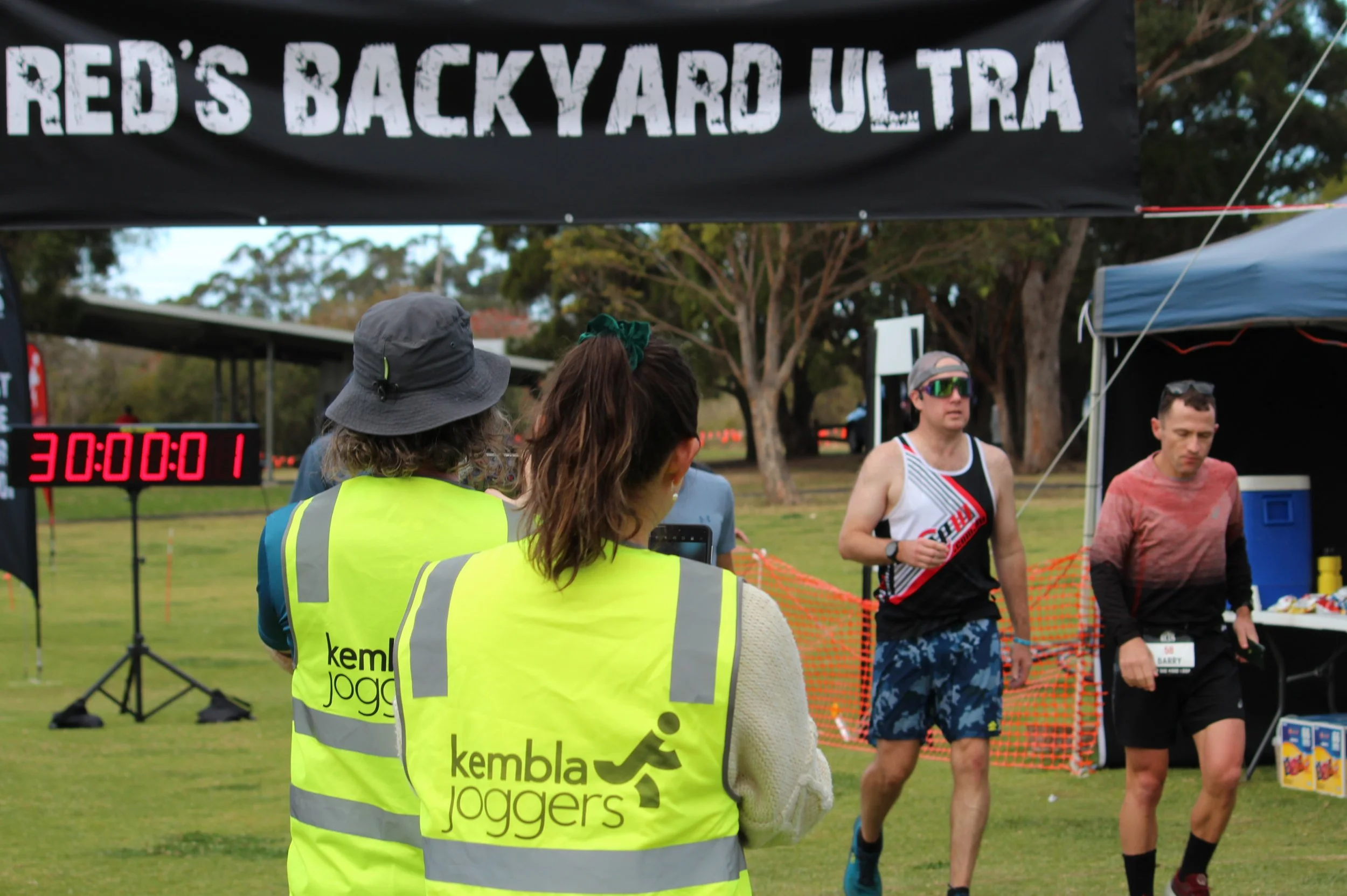 Two volunteers wearing high-visibility vests with 'Kembla Joggers' logo watch runners finish the race at the 'Red's Backyard Ultra' event. Runners approach the finish line, with a digital timer reading 30:00:01, and a black banner overhead displays e