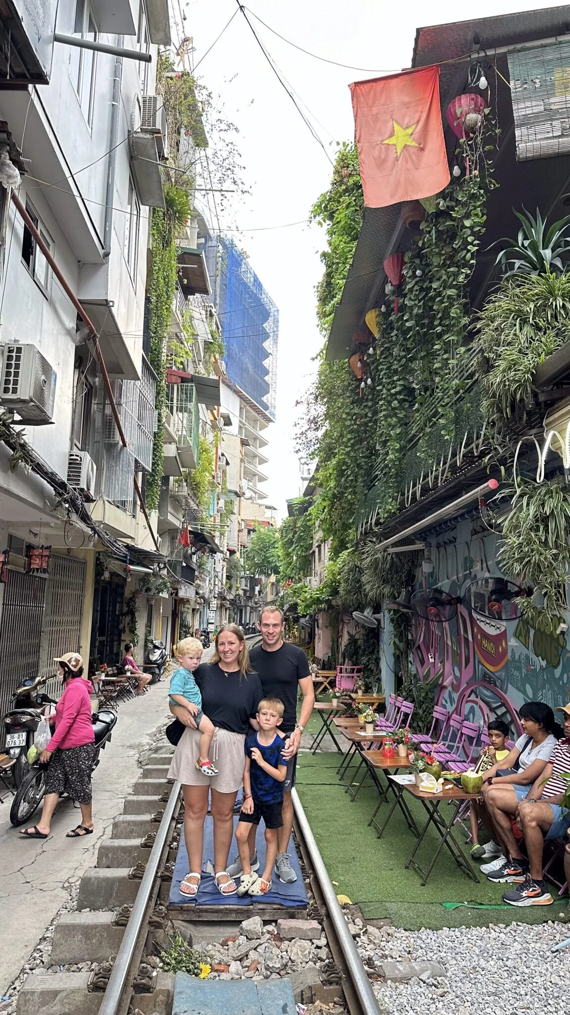 A family of four standing on train tracks in an urban alleyway, with people sitting at outdoor tables on the right and buildings with hanging plants and flags on the left.