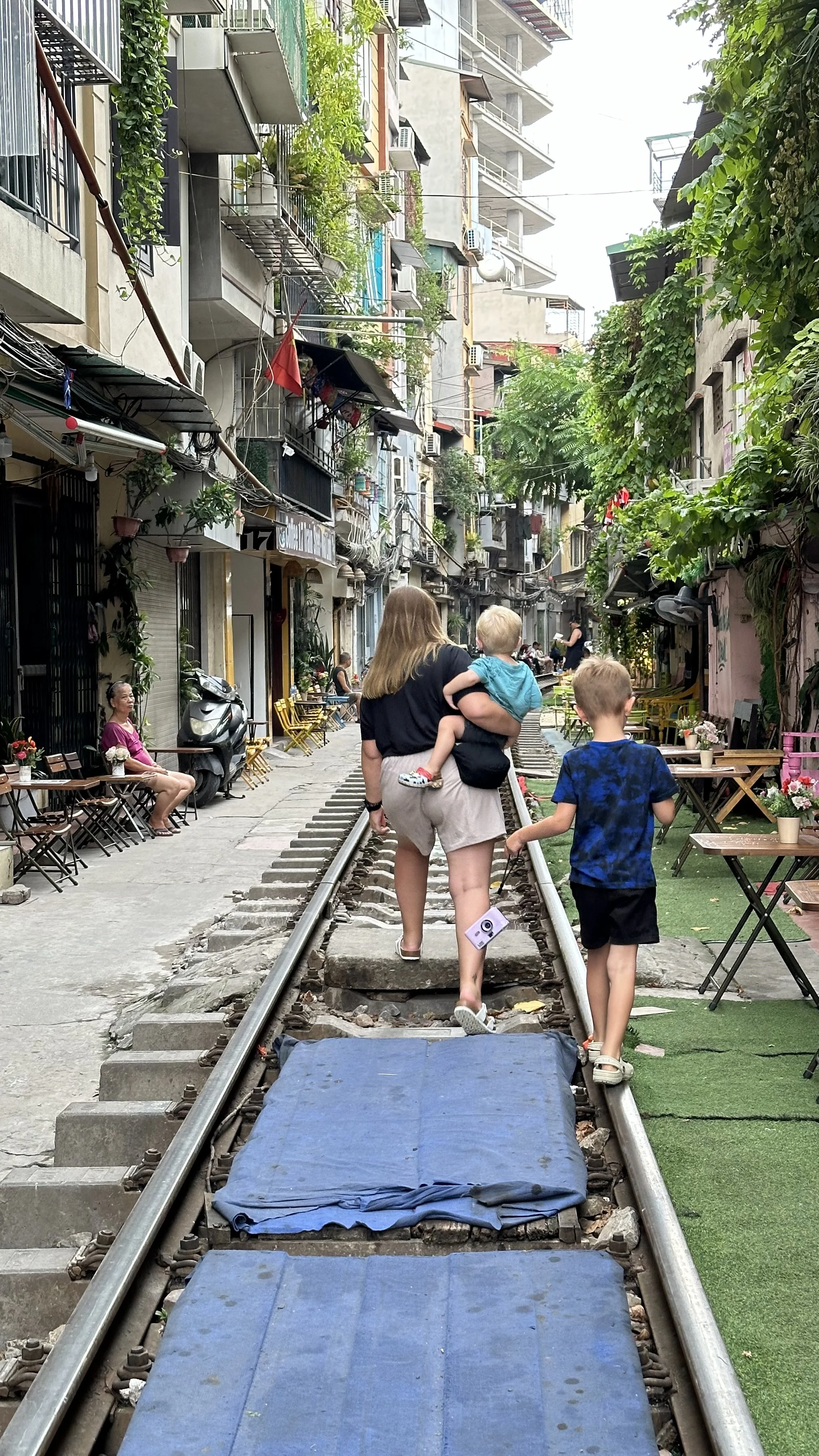 A woman and two young boys walk along a railway track that runs through a narrow street in a busy urban area. The street is lined with small shops, cafes, and multi-story residential buildings with balconies and greenery. People sit outside cafes, flowers decorate the area, and the scene has a lively, eclectic atmosphere.