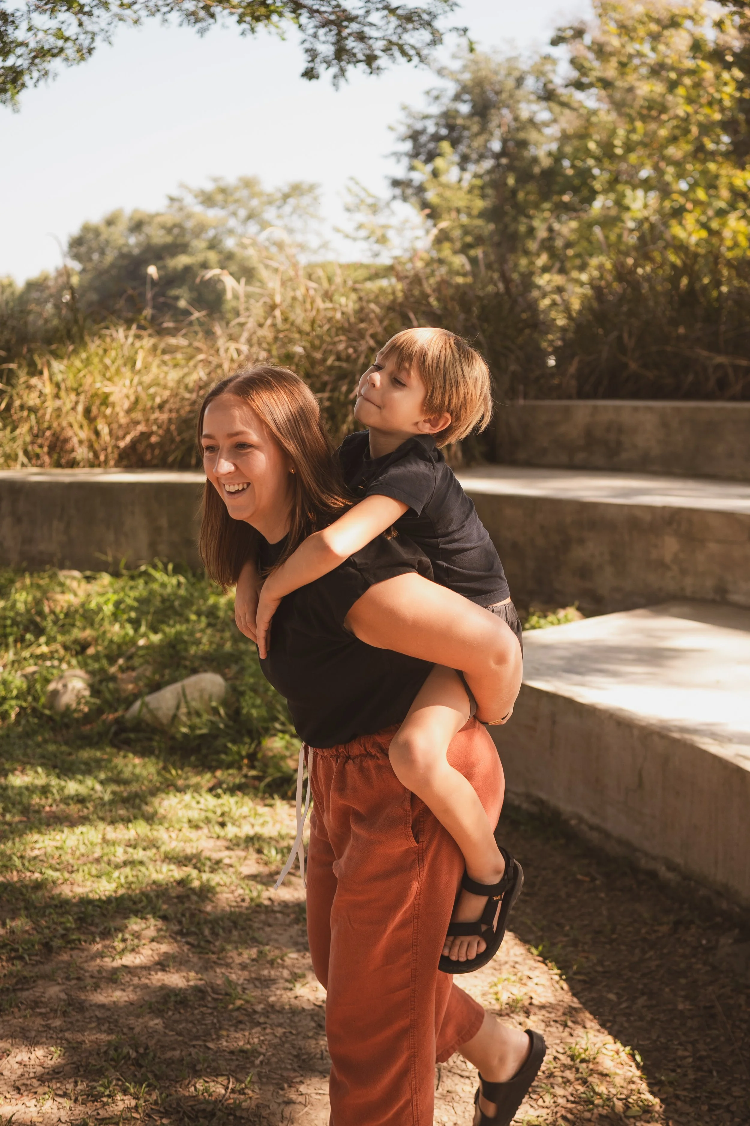 A woman giving a piggyback ride to a smiling young boy outdoors on a sunny day, with greenery and steps in the background.