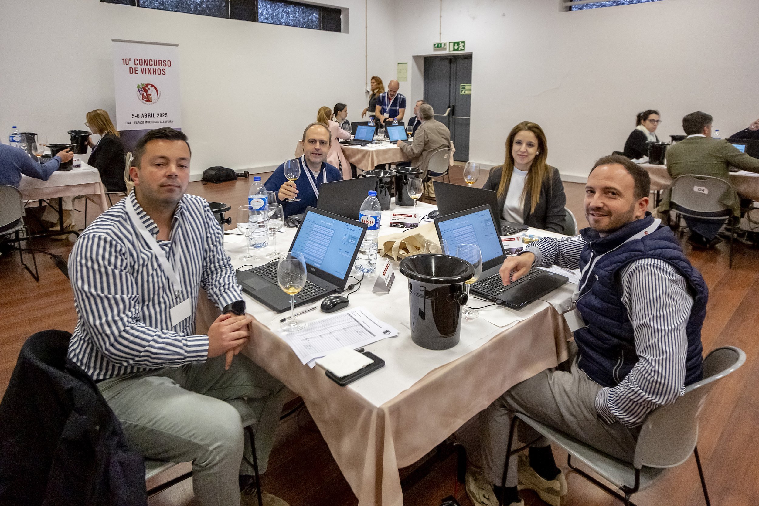 Participantes em uma conferência de vinhos em uma sala de eventos, sentados ao redor de uma mesa com laptops, garrafas de água, taças de vinho, papéis e aparelhos eletrônicos.