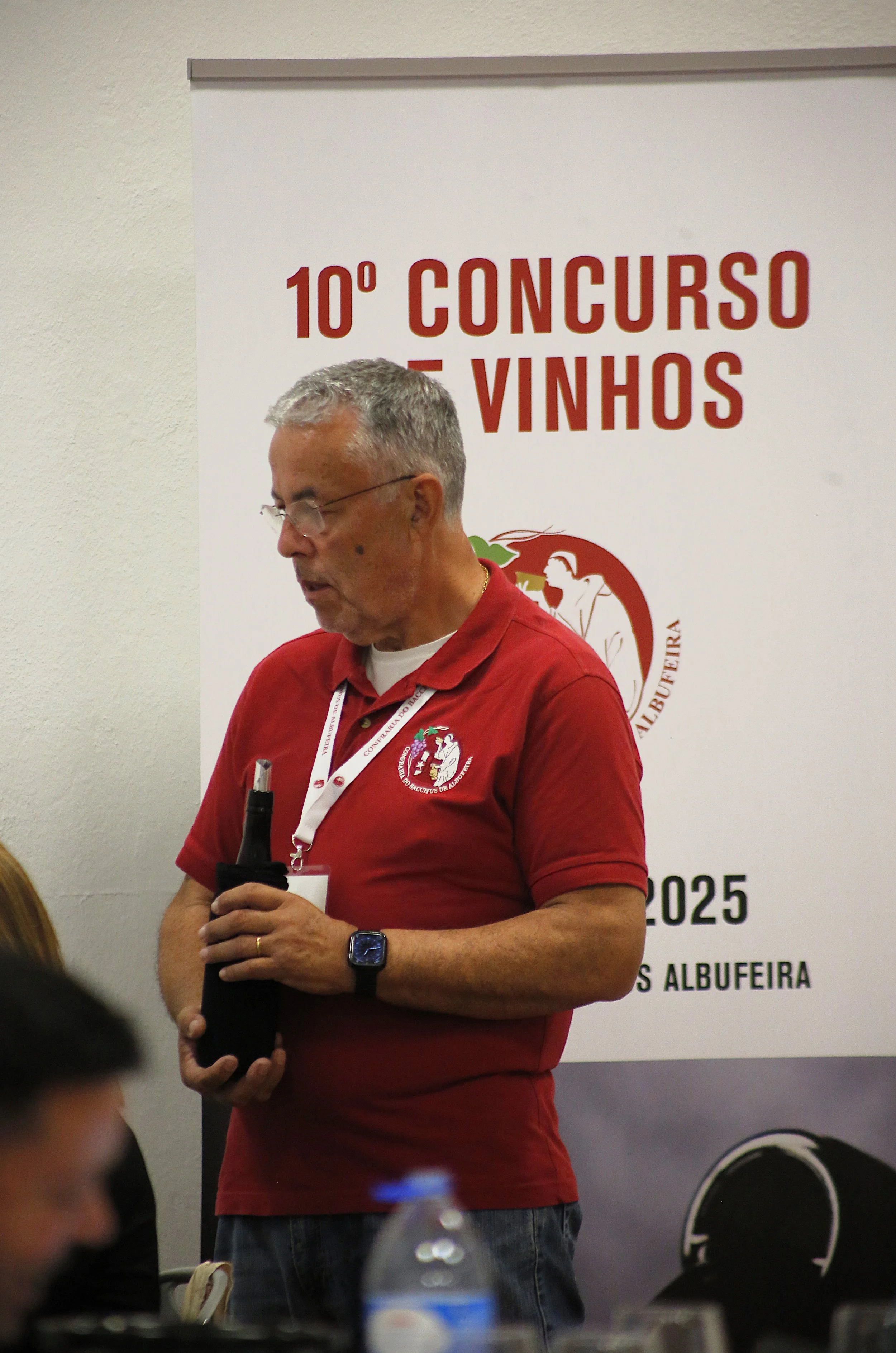 Homem segurando uma garrafa de vinho em frente a placa de 10º Concurso de Vinhos, com uma pessoa parcialmente visível ao lado, em evento de vinhos em Albufeira.