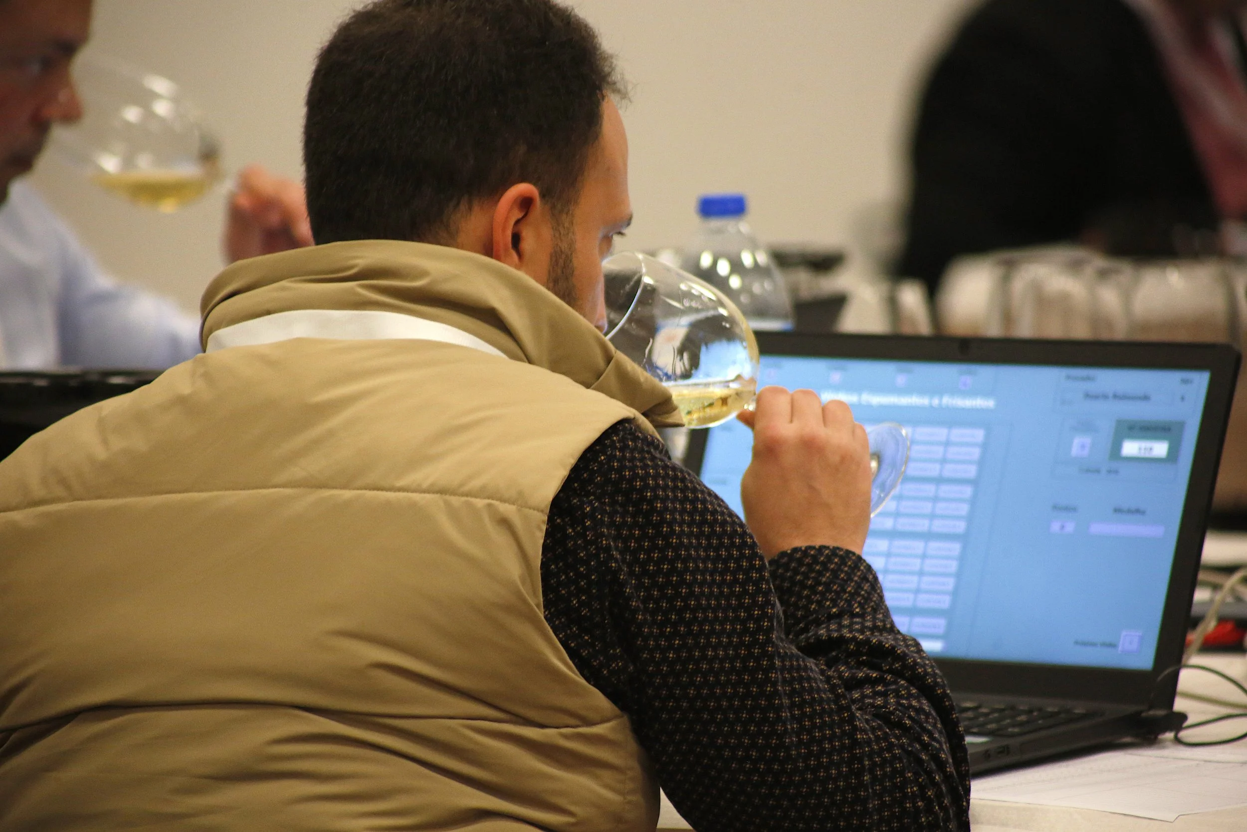 Homem assistindo a uma apresentação ou reunião, bebendo vinho, sentado diante de um computador portátil em uma sala de conferências.