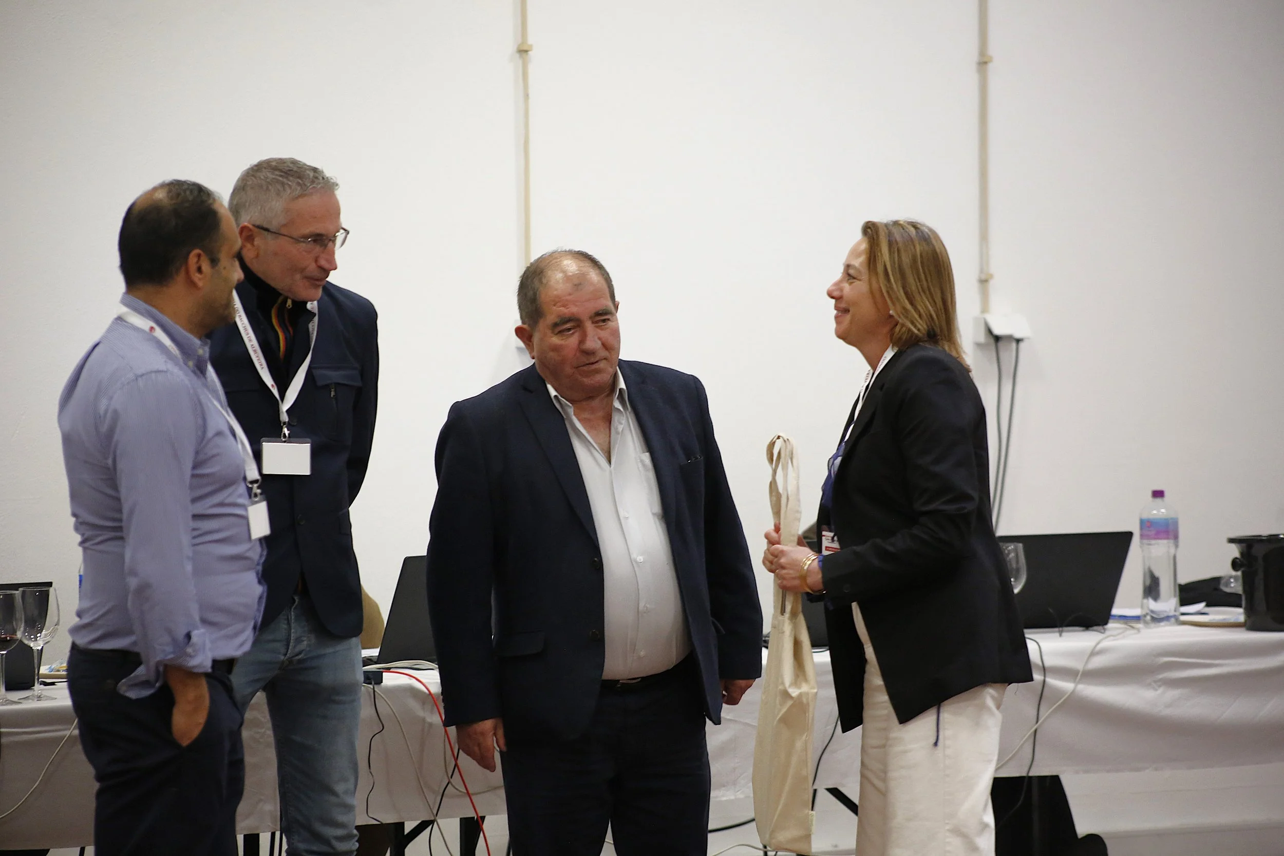 Quatro pessoas em uma sala de reunião, algumas com crachás, conversando, uma mulher entrega uma sacola a um homem. Mesas com laptops, garrafas de água e objetos de escritório ao fundo.