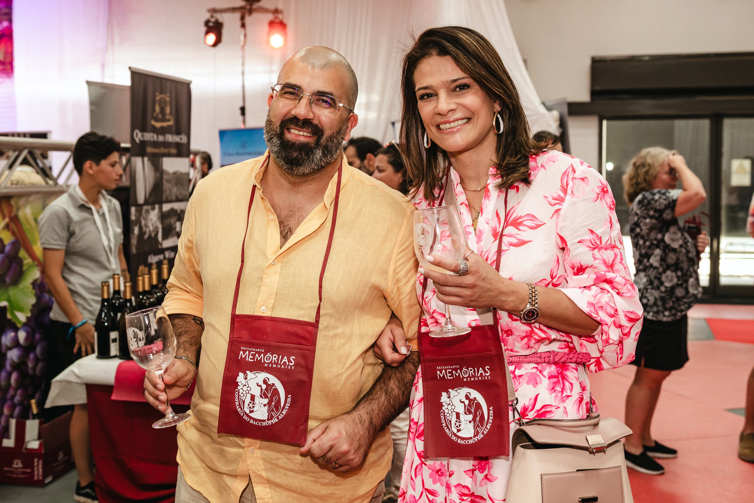 Homens e mulheres sorrindo segurando taças de vinho em um evento social, com banner e garrafas de vinho ao fundo.