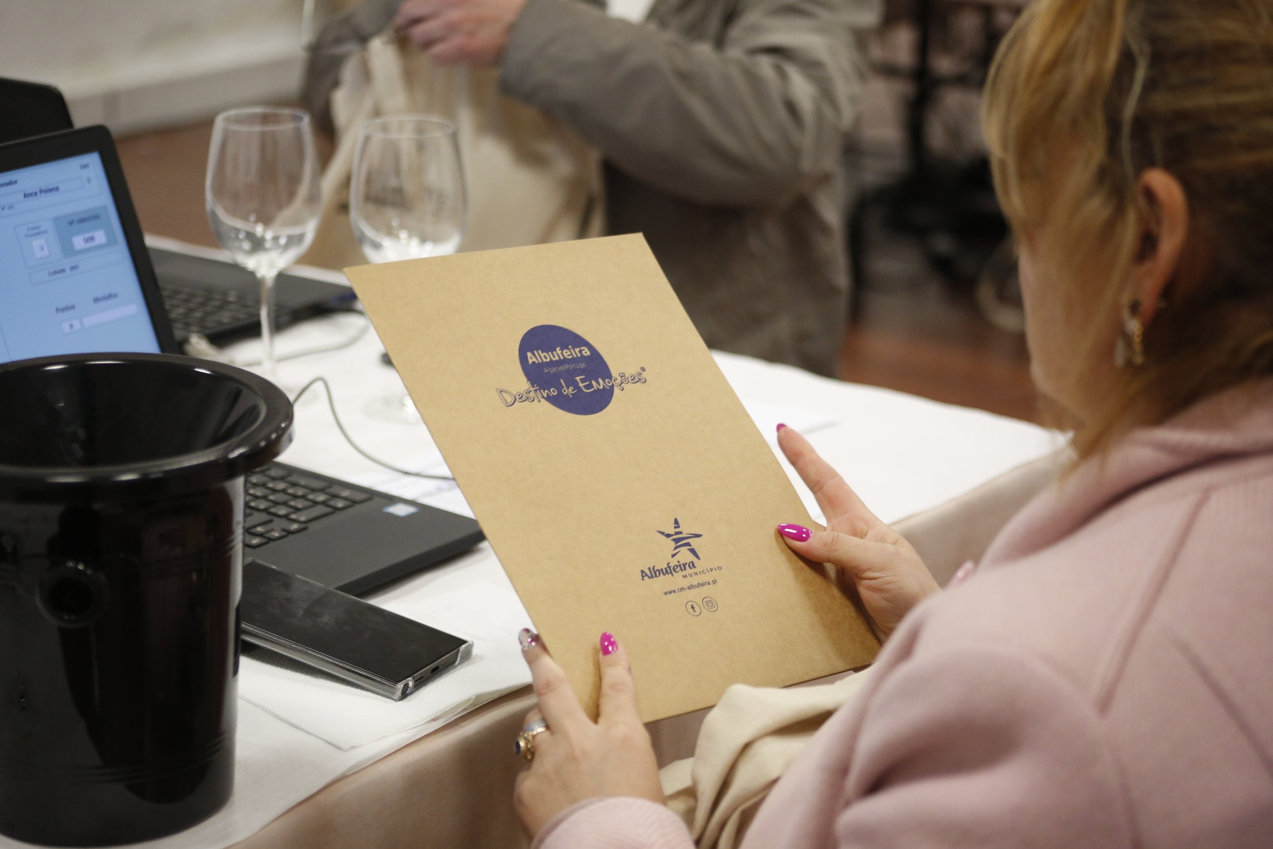 Mulher lendo um folder de papel com logotipos e textos relacionados à Albufeira, em uma mesa com laptop, copos de água e outros objetos.