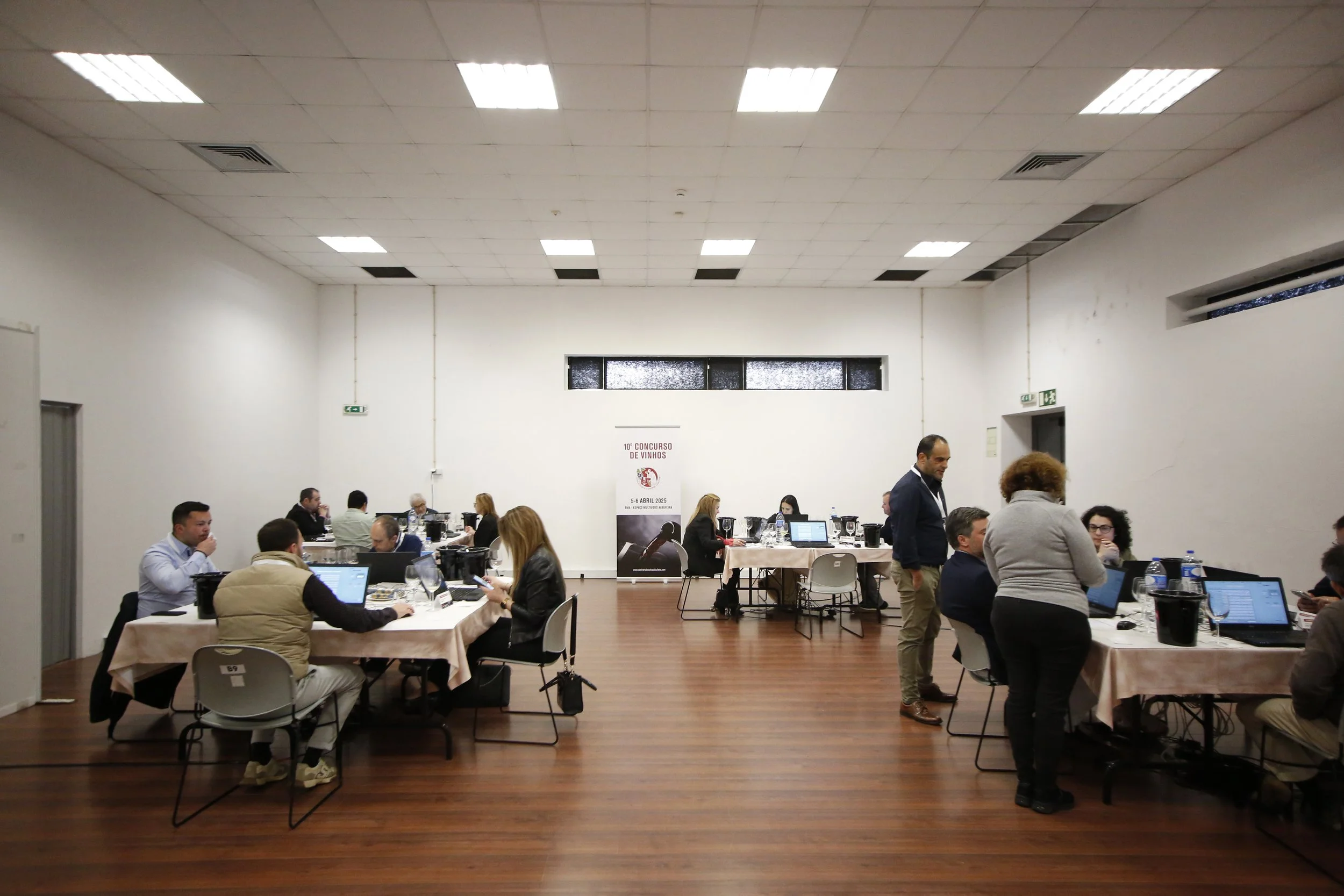 Sala de eventos com várias pessoas sentadas em mesas, trabalhando com laptops, em um concurso de vinhos. Há uma parede branca ao fundo com um banner que diz '10º Concurso de Vinhos'.