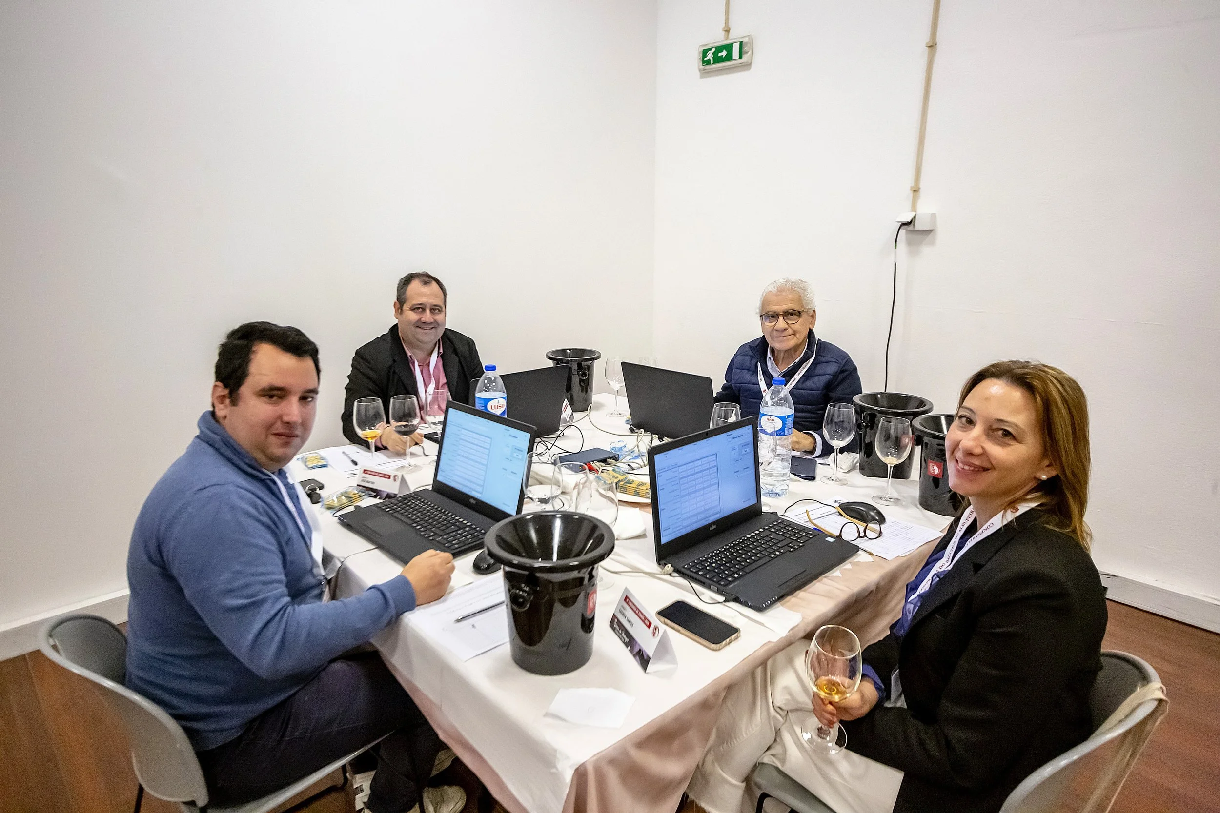 Grupo de cinco pessoas sentadas ao redor de uma mesa em uma reunião, com laptops, garrafas de água, taças de vinho e papéis, em um ambiente interno com paredes brancas e piso de madeira.