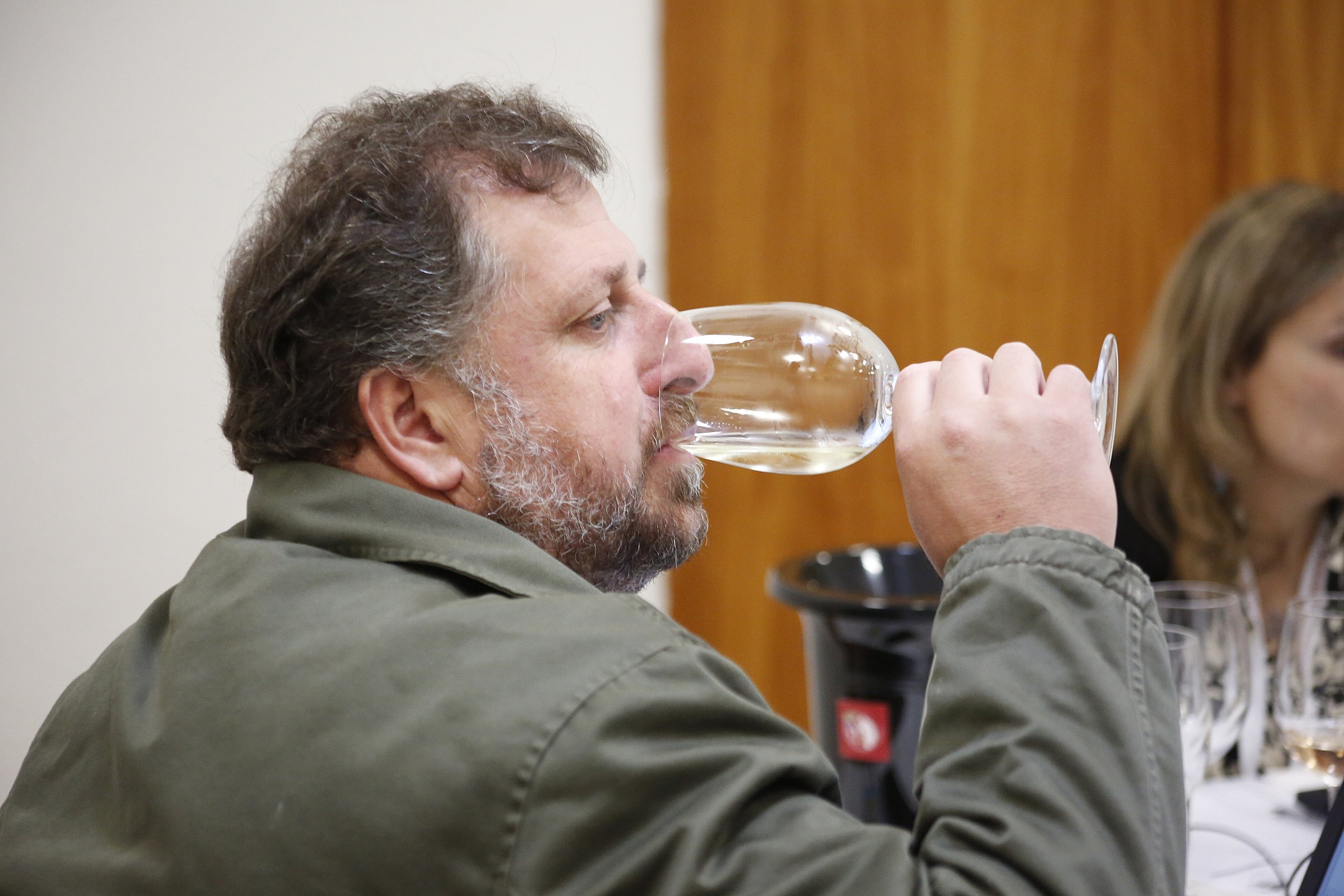 Homem com barba e cabelo grisalho, vestindo jaqueta cinza, bebendo champanhe em taça durante uma reunião ou evento social.