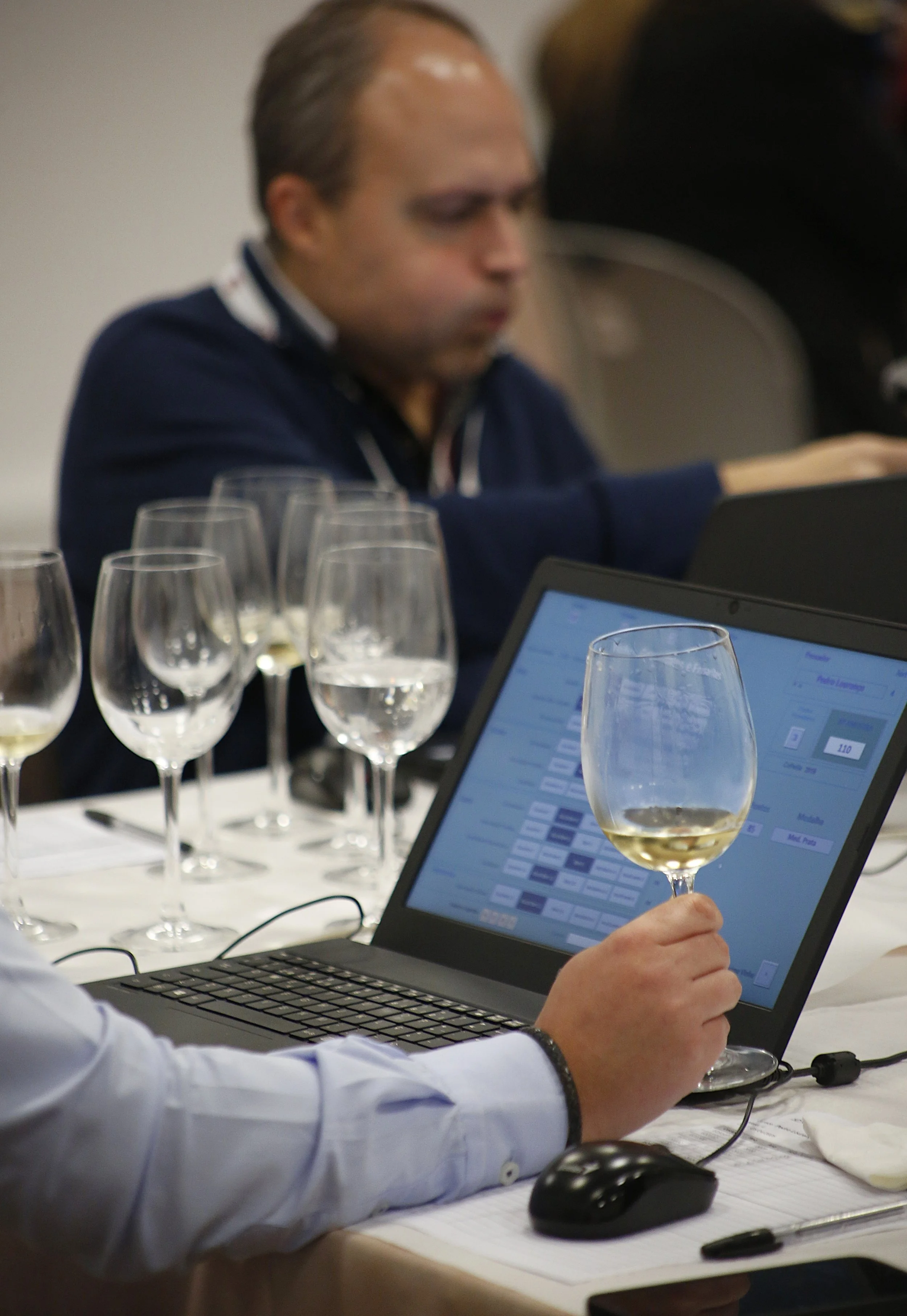 Pessoa manuseando taça de vinho em frente a um laptop em uma mesa de apresentação, com várias taças de vinho ao redor e uma pessoa ao fundo.