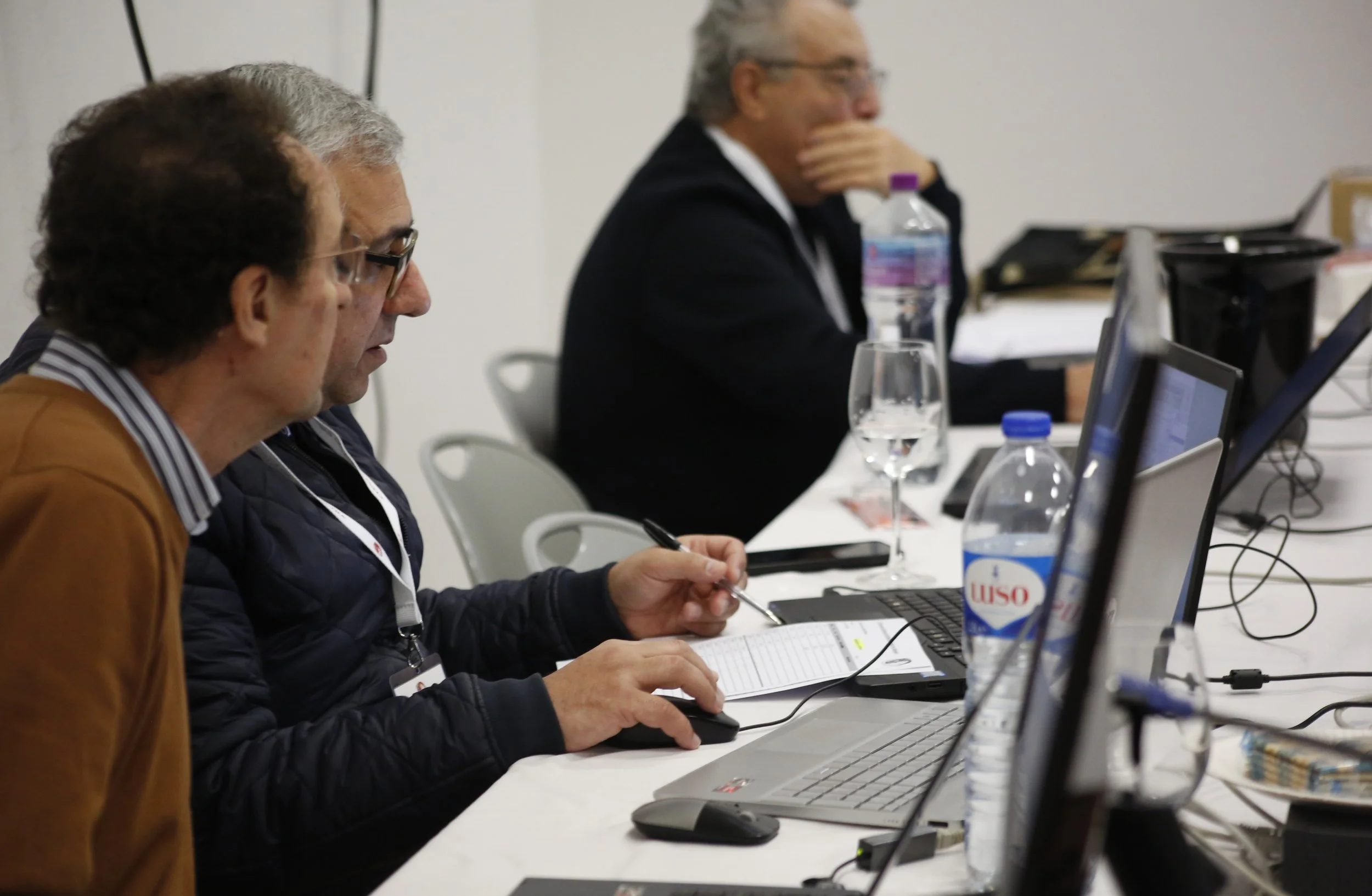 Pessoas em uma reunião de trabalho, usando laptops e anotando informações, com garrafas de água na mesa.