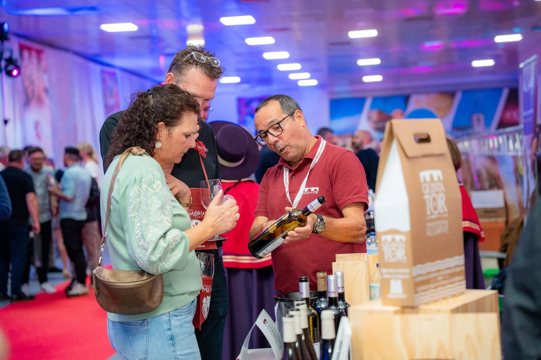 Homens e mulher conversando na feira de vinhos, com vários barris e garrafas expostos.