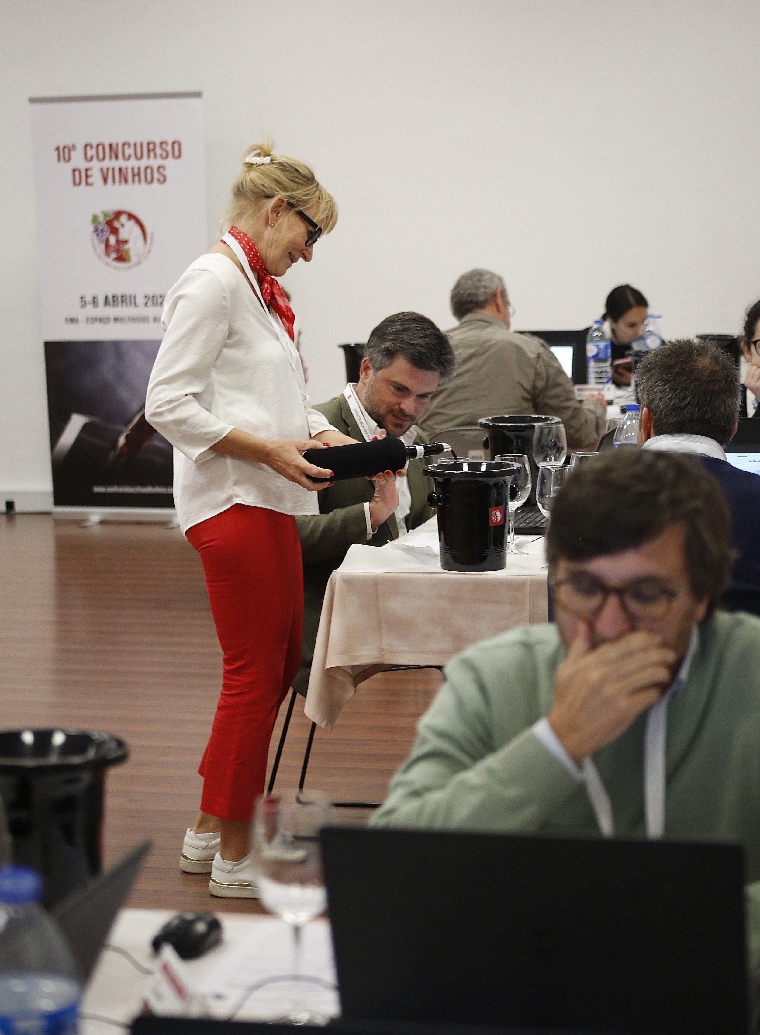 Pessoas em uma sala de evento do concurso de vinhos, com uma mulher servindo vinho para um homem sentado à mesa. Em segundo plano, outros participantes usando laptops.