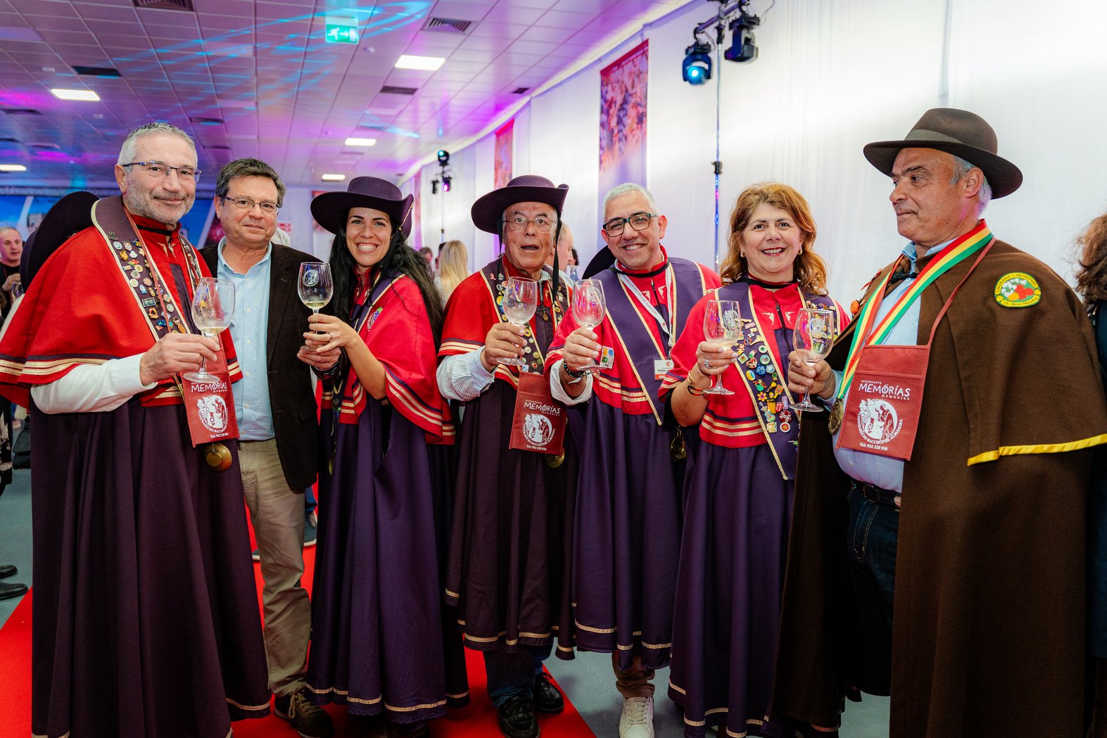 Grupo de pessoas em traje tradicional, segurando taças de vinho, em ambiente festivo com iluminação colorida, decoração com bandeiras, participando de um evento cultural ou comemorativo.