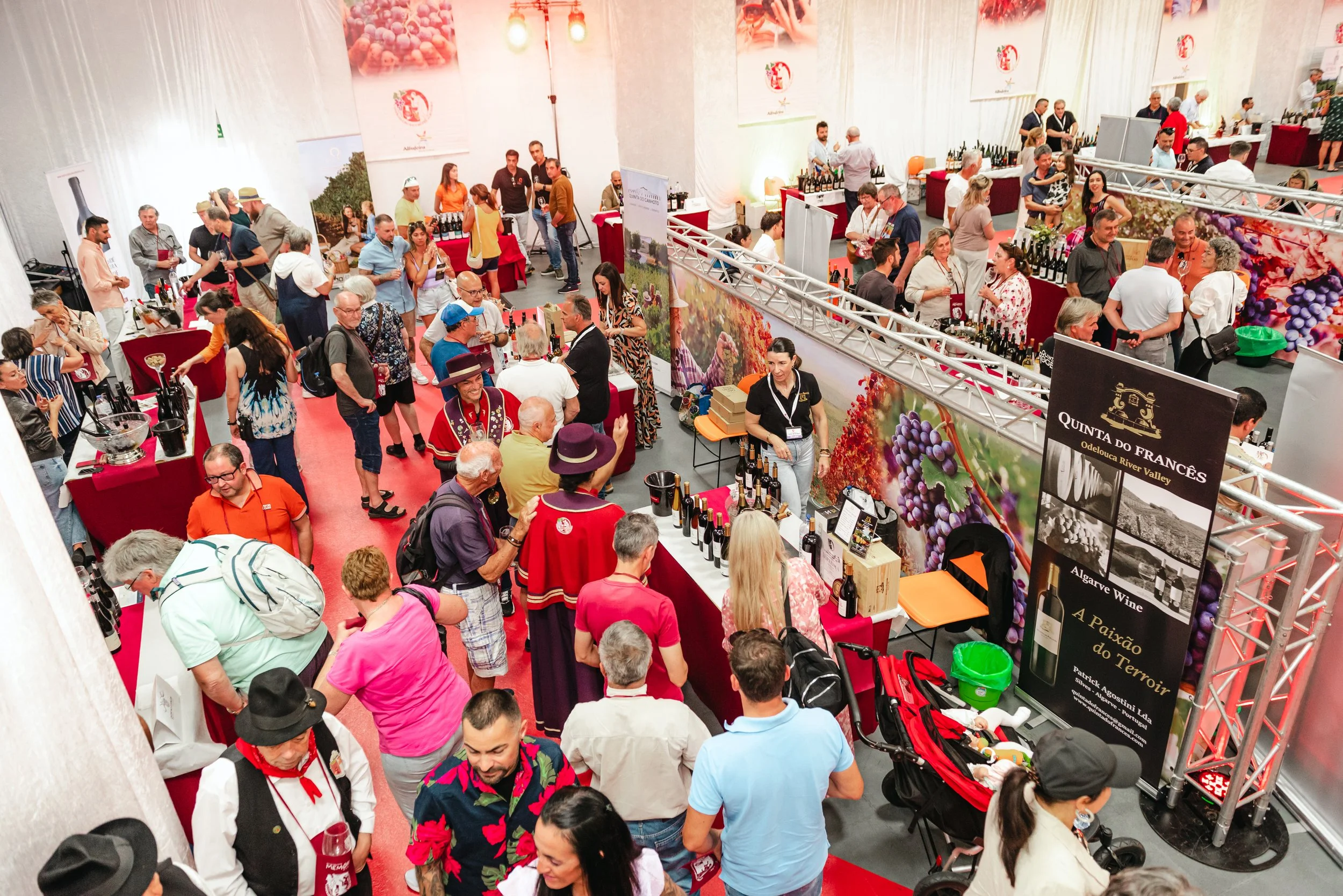 Evento de degustação de vinhos com muitas pessoas explorando diferentes barracas, algumas vestidas com roupas tradicionais, e banners de vinícolas portugueses, como Quinta do Francês. Decoração vibrante com imagens de uvas e vinícolas.