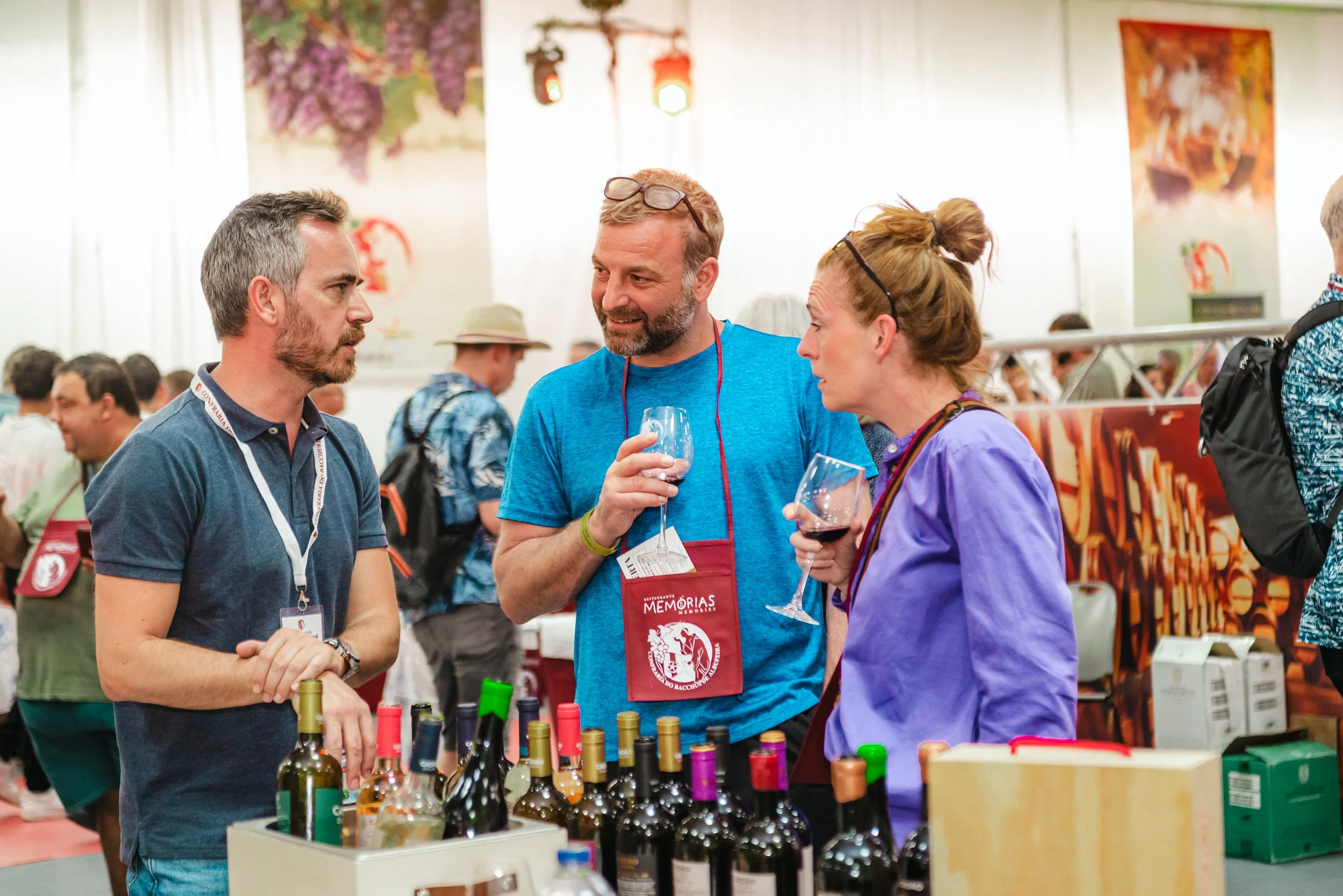 Três pessoas conversando em um evento de vinhos, segurando taças com vinho, ao lado de uma mesa com várias garrafas de vinho. Pessoas ao fundo também estão no evento, que parece ser uma feira ou feira de vinhos.