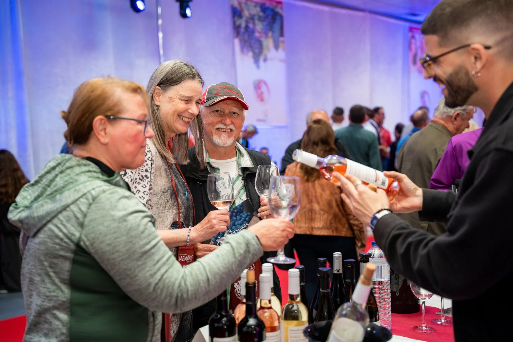 Pessoas sorridentes em uma feira de vinhos, segurando taças de vinho rosé, enquanto um vendedor apresenta uma garrafa de vinho. Mesas com garrafas de vinho ao fundo, ambiente animado e colorido.