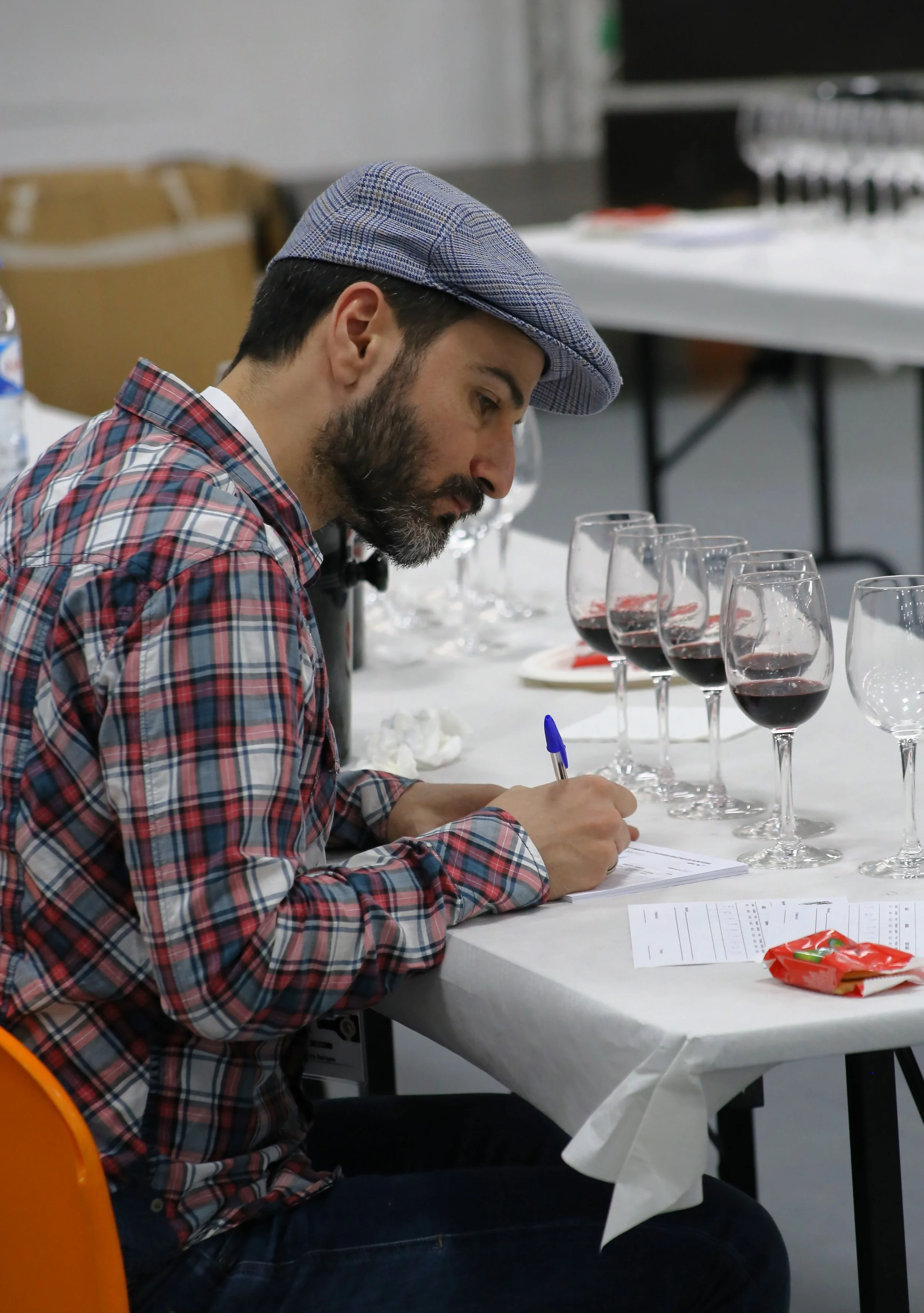 Homem com barba, vestindo camisa xadrez e boina, senta-se à mesa com taças de vinho tinto e faz anotações em uma folha de papel.