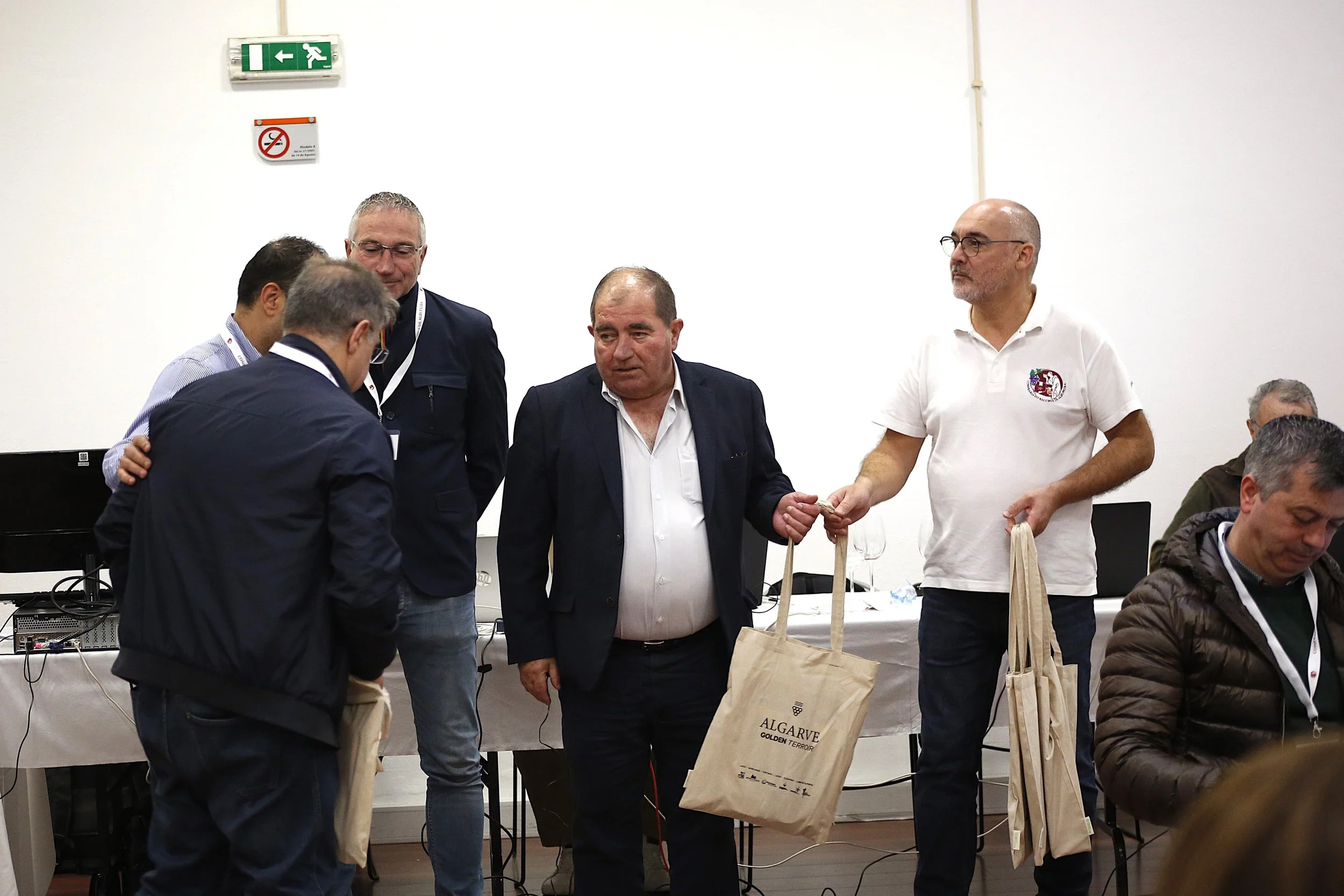 Homens trocando brindes ou presentes em uma sala com mesa e computadores ao fundo, alguns usando crachás e roupas formais, na conferência ou evento.