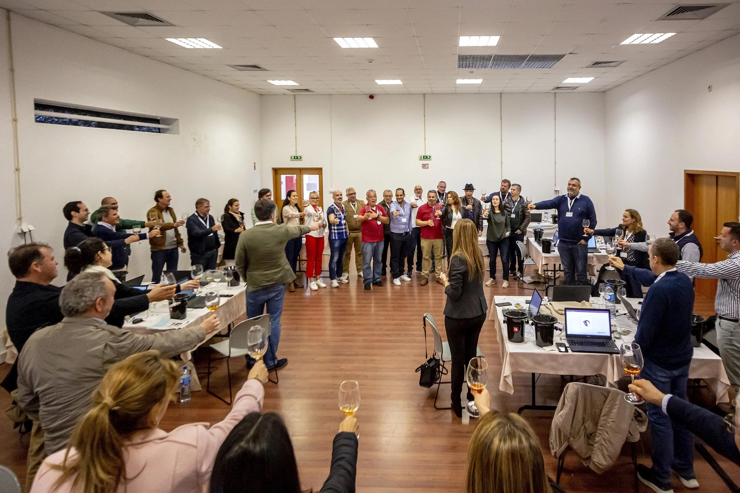 Grupo de pessoas em uma reunião ou evento, formando um círculo e brindando com taças de vinho em uma sala de conferências com mesas, laptops e equipamentos de áudio.