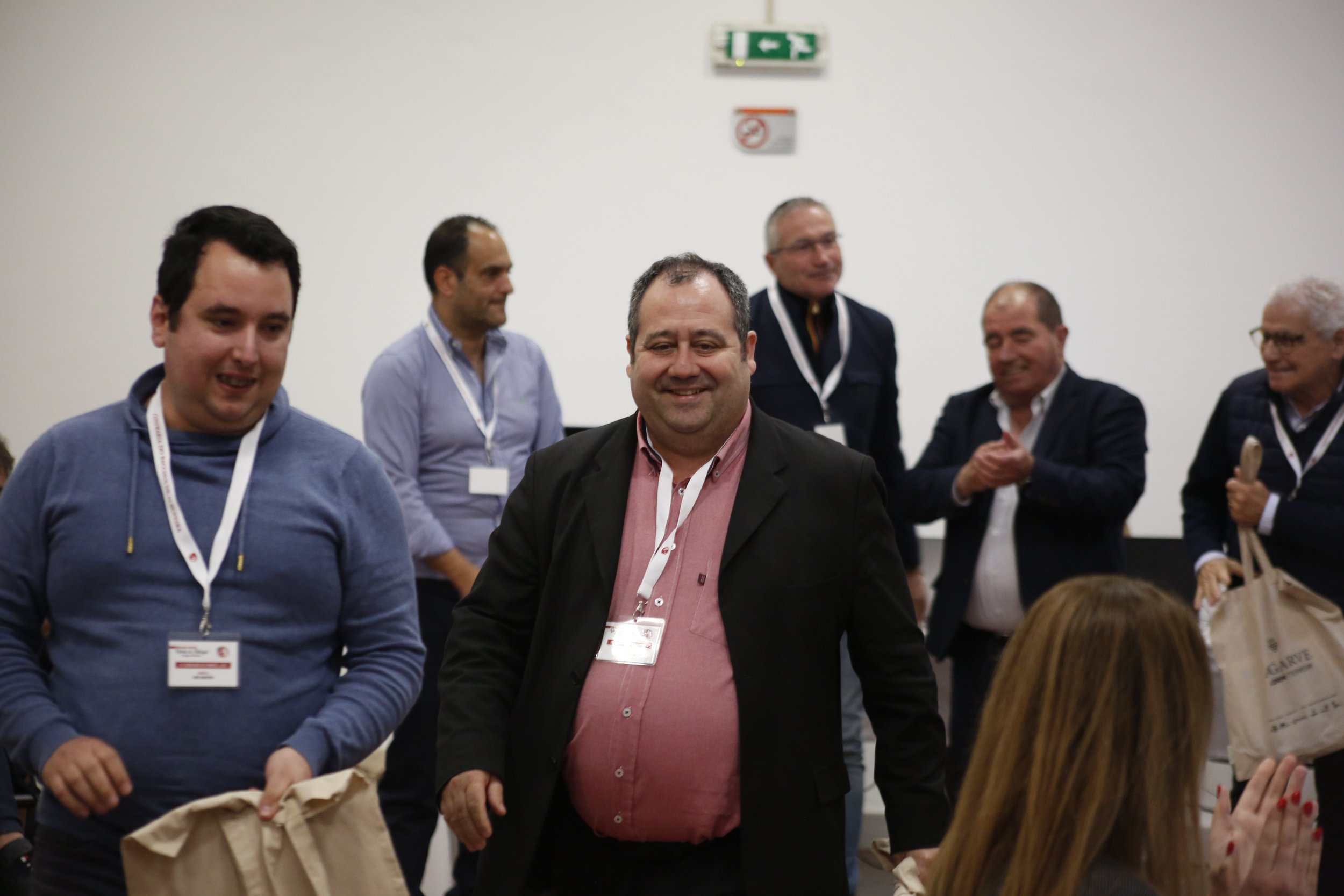 Grupo de pessoas sorrindo e conversando em um ambiente de conferência ou reunião, com crachás de identificação ao pescoço.