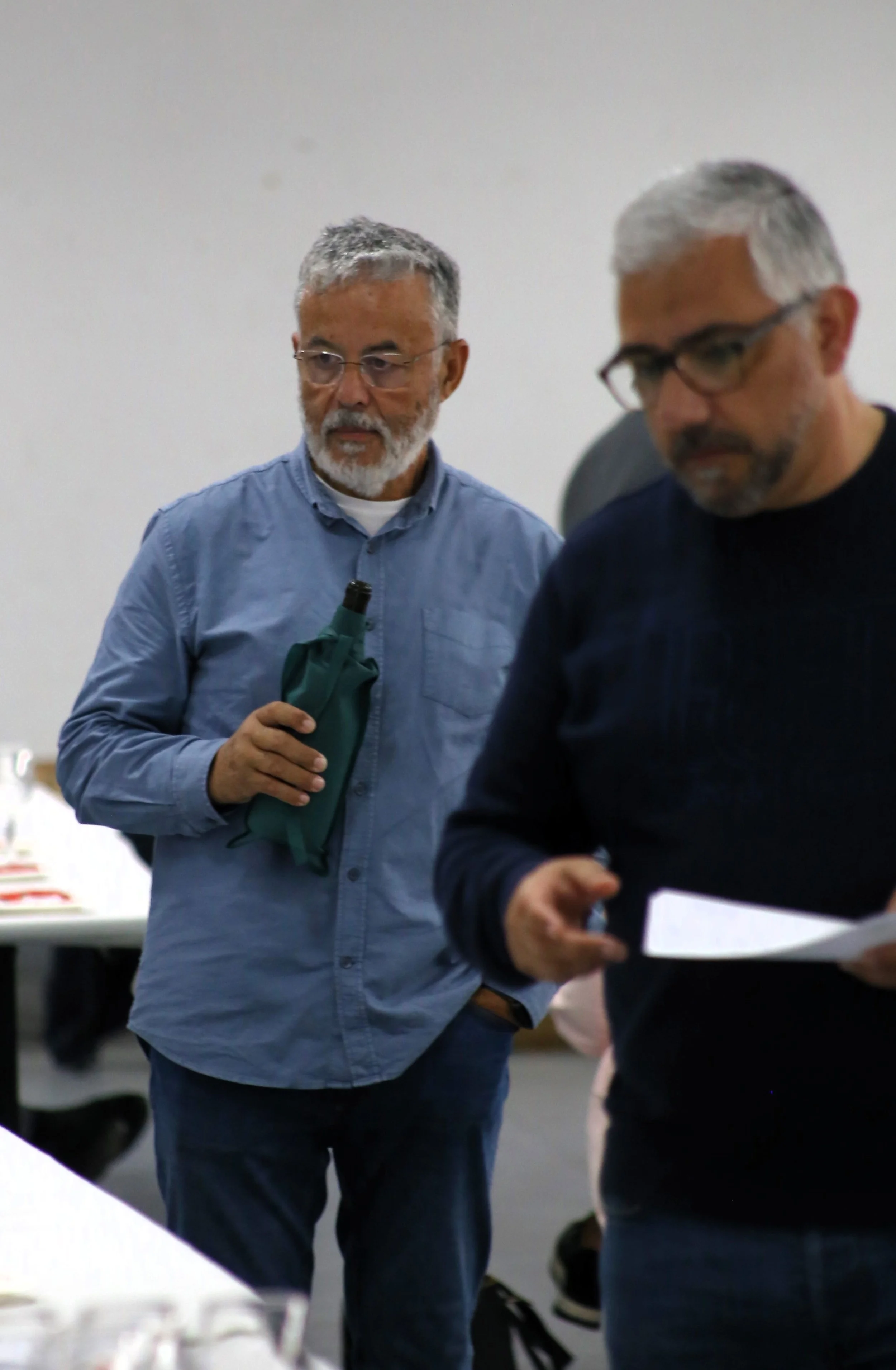 Homem com barba e óculos segurando uma garrafa e observando uma folha de papel, ao lado de outro homem que também está segurando uma folha de papel, em ambiente interno