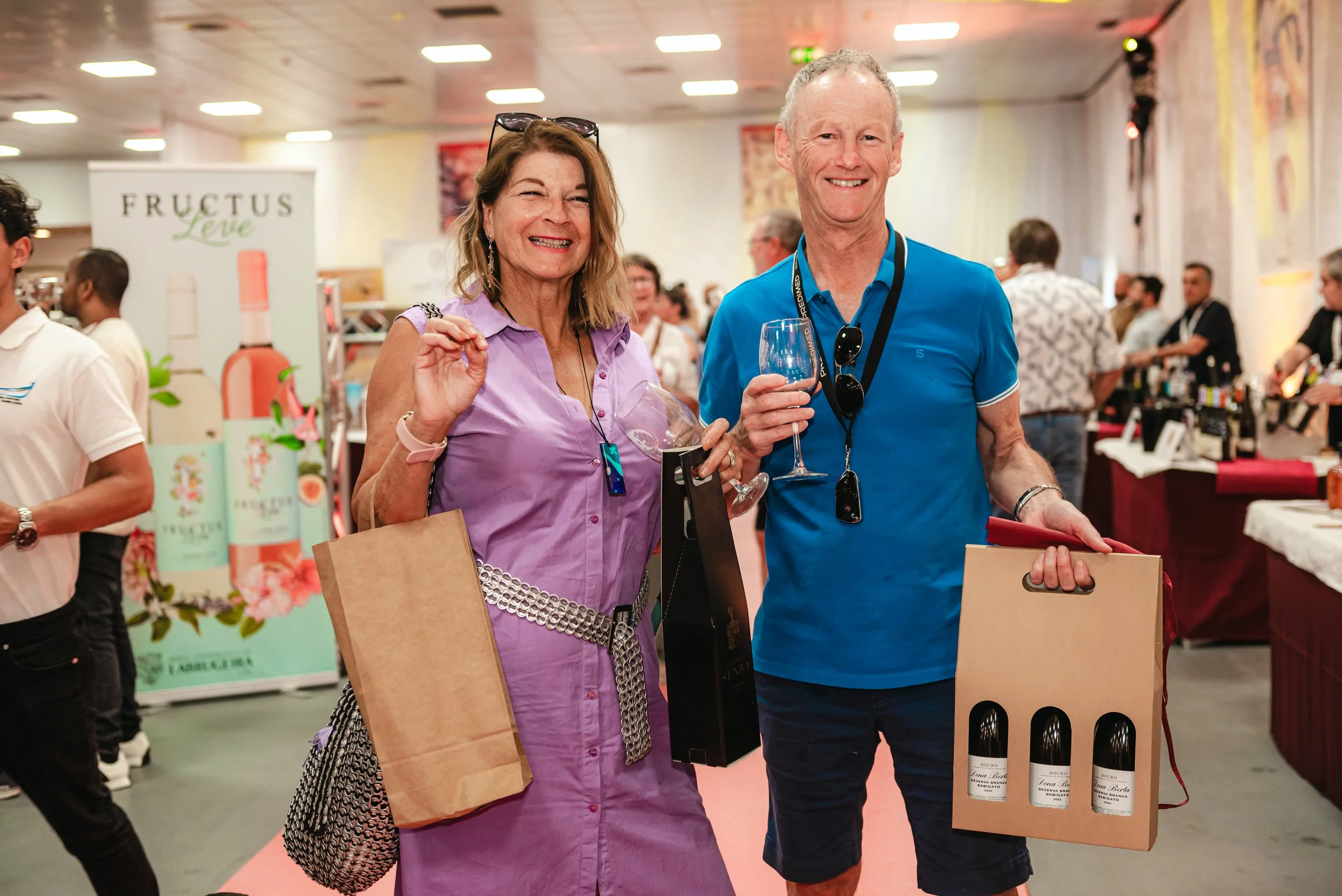 Homens e mulheres sorrindo em feira de vinhos, segurando taças e caixas de vinhos.