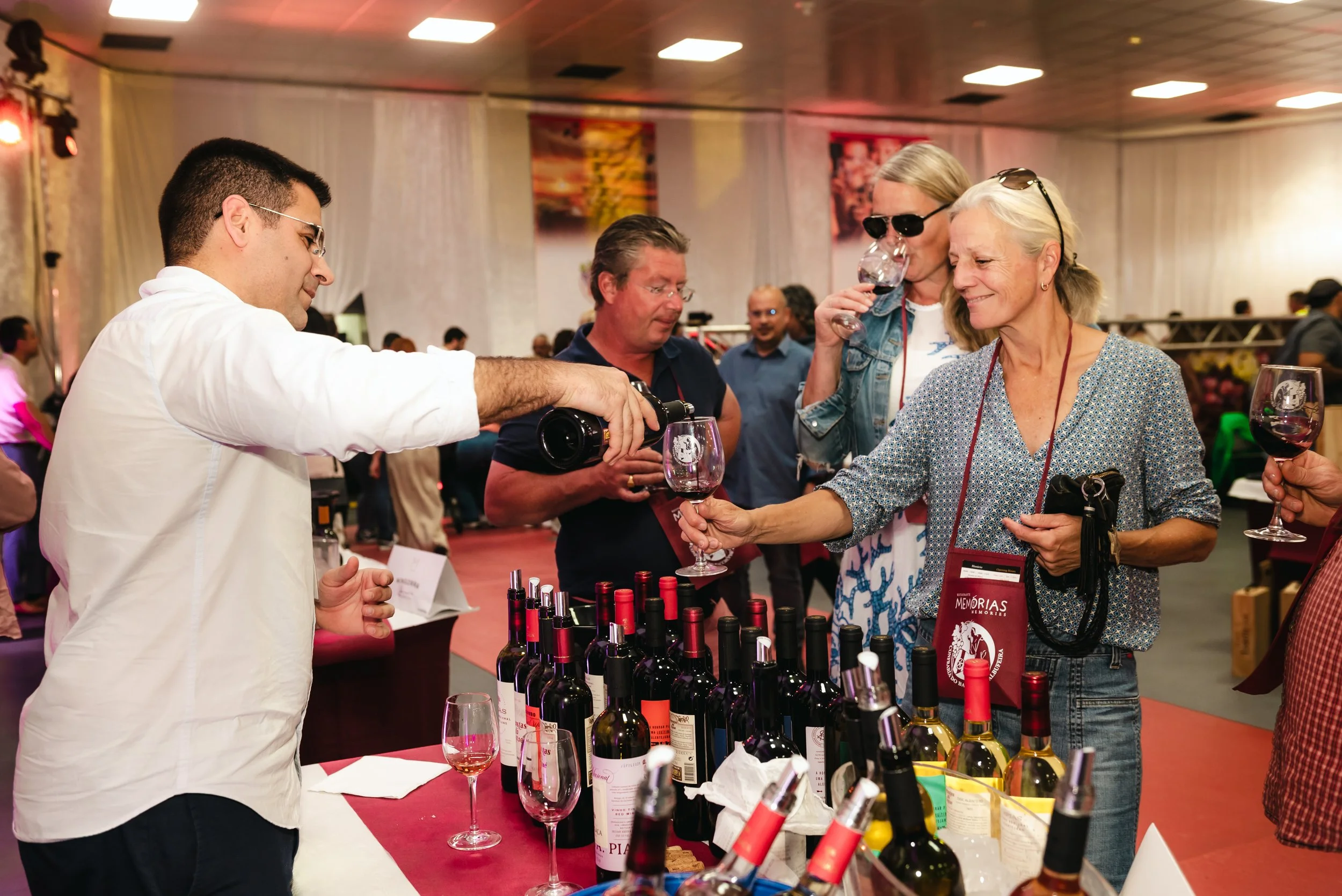 Pessoas degustando vinho na feira de vinhos, com diversas garrafas na mesa e várias pessoas ao fundo