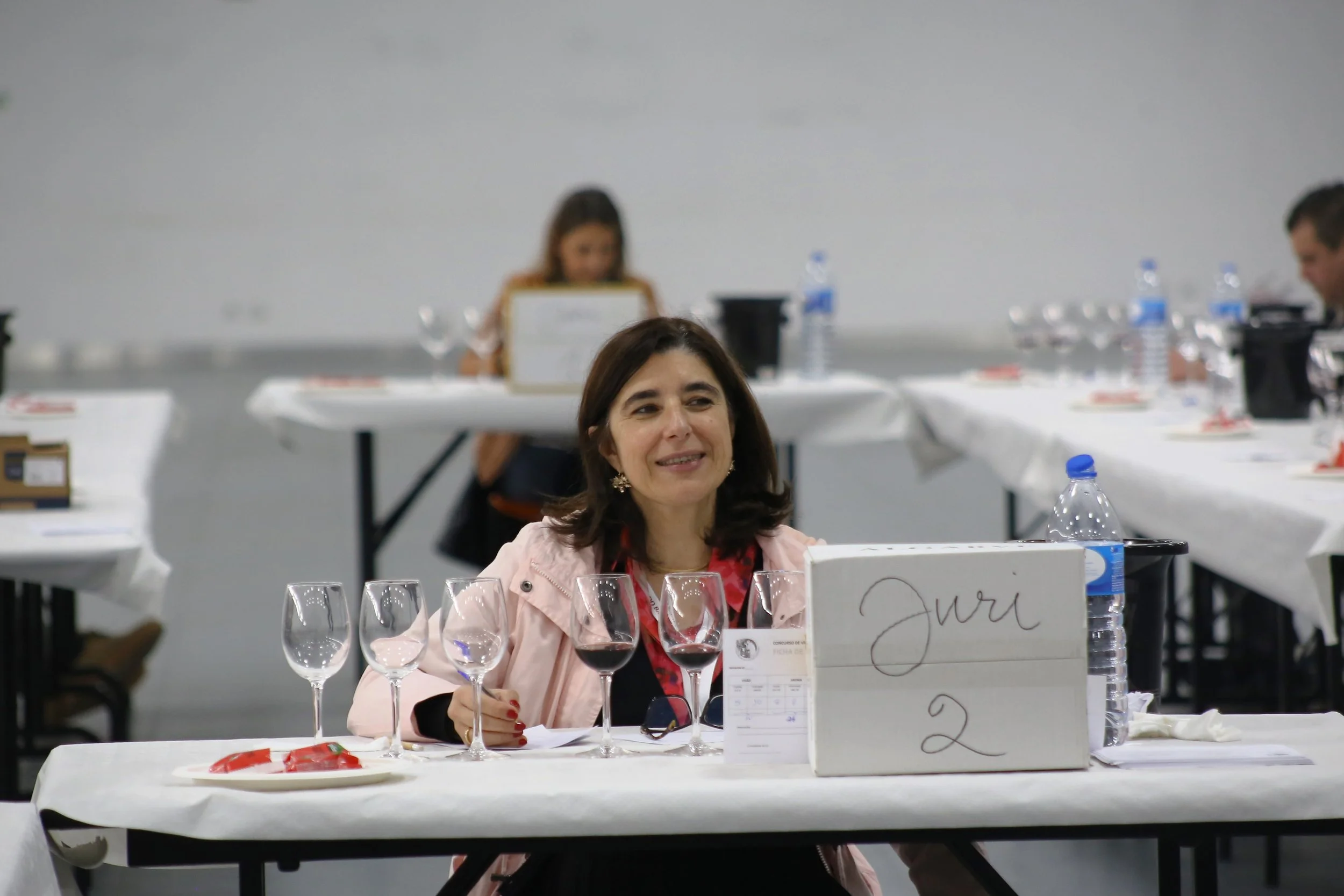 Mulher sorridente sentada em uma mesa de degustação de vinhos com três taças de vinho, uma de cada cor (branco, rosé e tinto), e uma placa com 'Juri 2'.