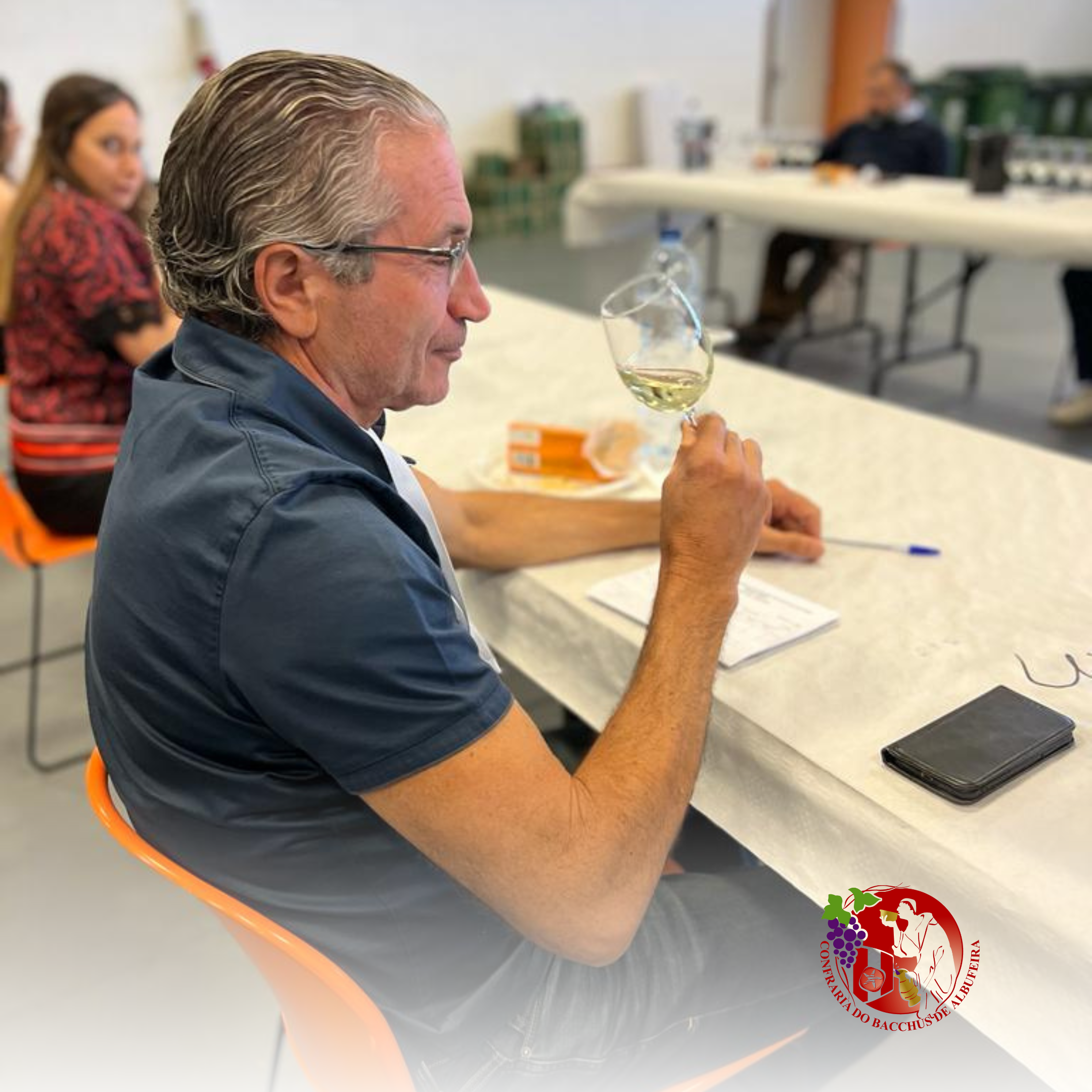 Homem com cabelo grisalho e óculos usando camiseta azul, sentado em uma mesa, segurando uma taça de vinho branco, com uma mulher ao fundo e objetos de papel e telefone na mesa. Logo de uma vinícola ou confeitaria na parte inferior direita.
