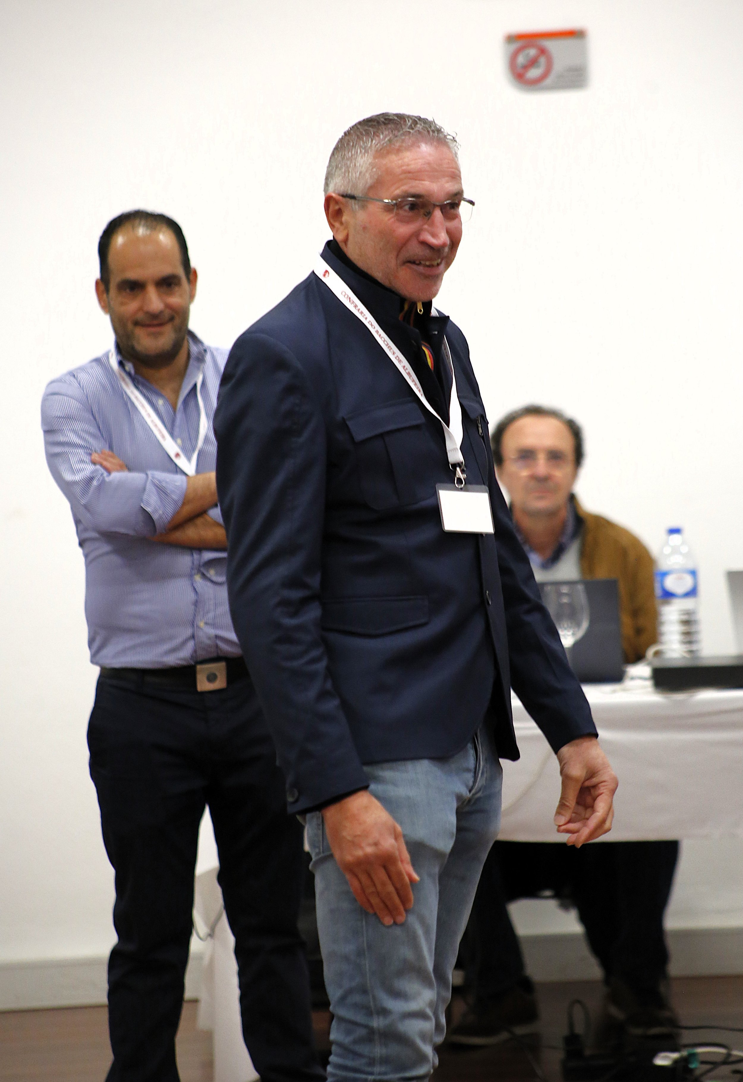 Homens em uma reunião ou conferência, com um deles sorrindo e vestindo um blazer azul escuro, enquanto outros estão ao fundo com computadores e garrafa de água na mesa.