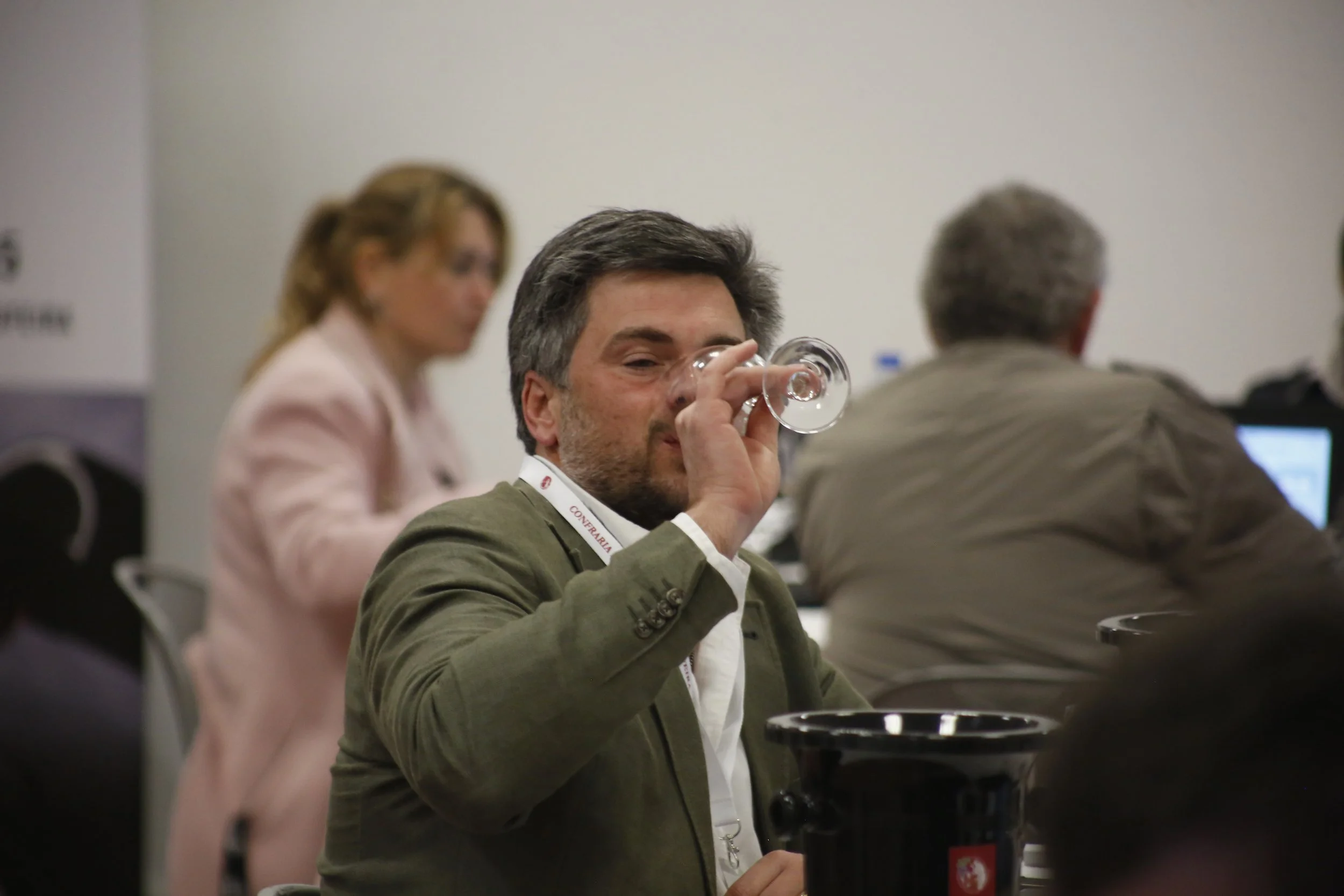 Homem tomando uma taça de vinho em uma conferência, com outras pessoas ao fundo usando computadores.