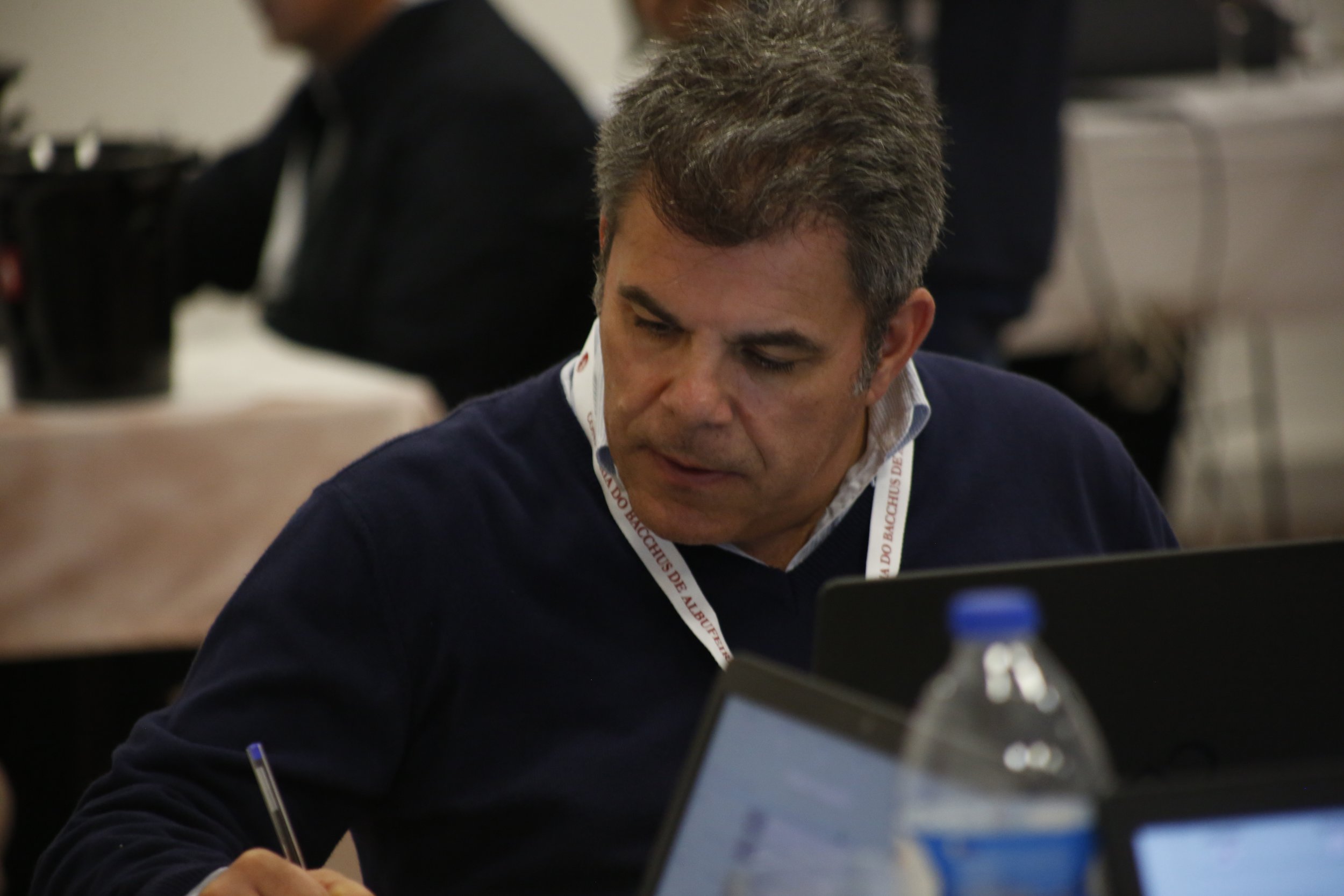 Homem de cabelo castanho escuro trabalhando com um computador portátil em uma sala de reunião, usando uma camisa branca e suéter azul escuro. Ao redor, outras pessoas também trabalham ou participam de reunião.