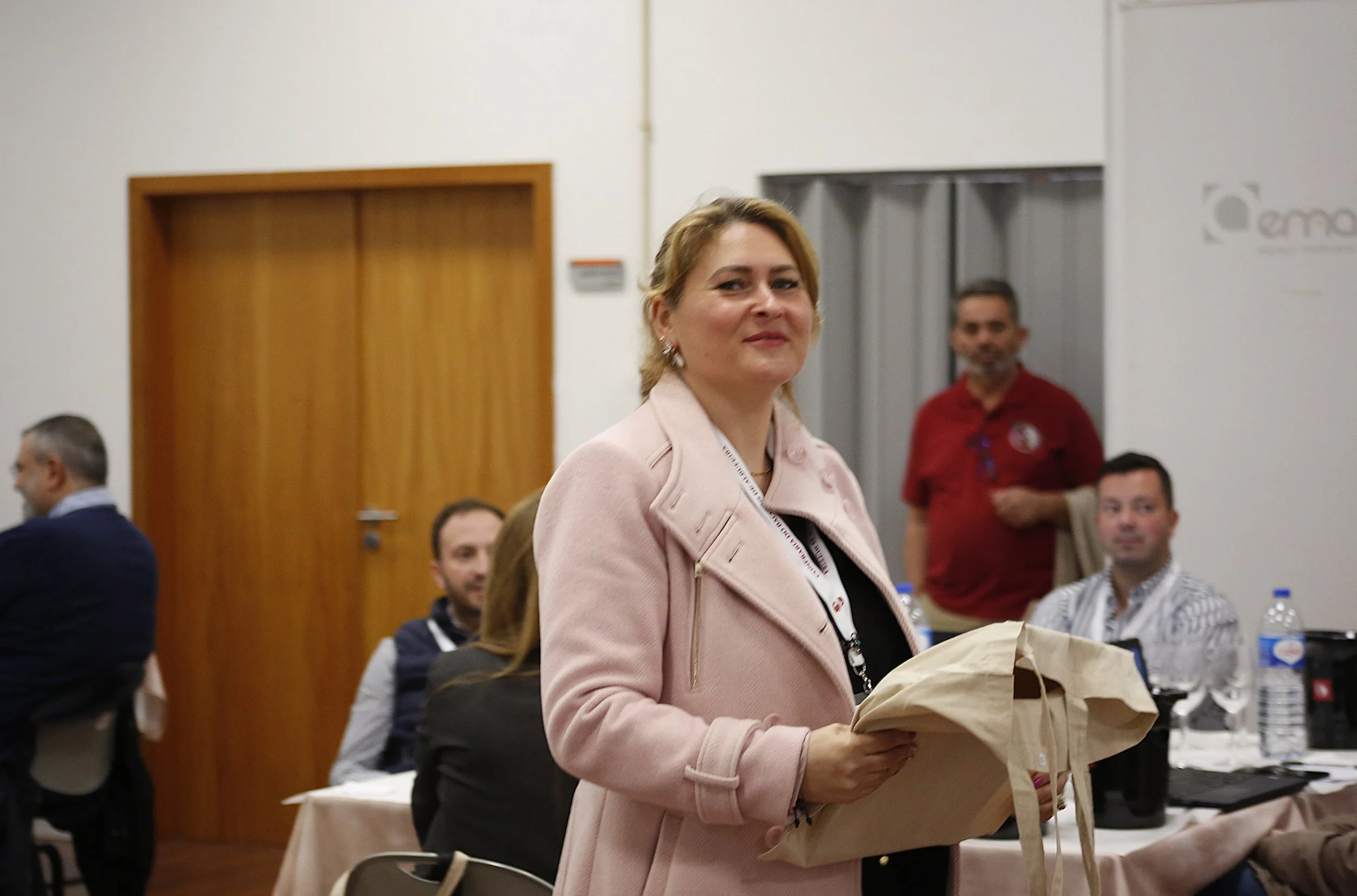 Mulher com blazer rosa segurando uma sacola de papel em uma sala de reunião com outras pessoas sentadas ao fundo.