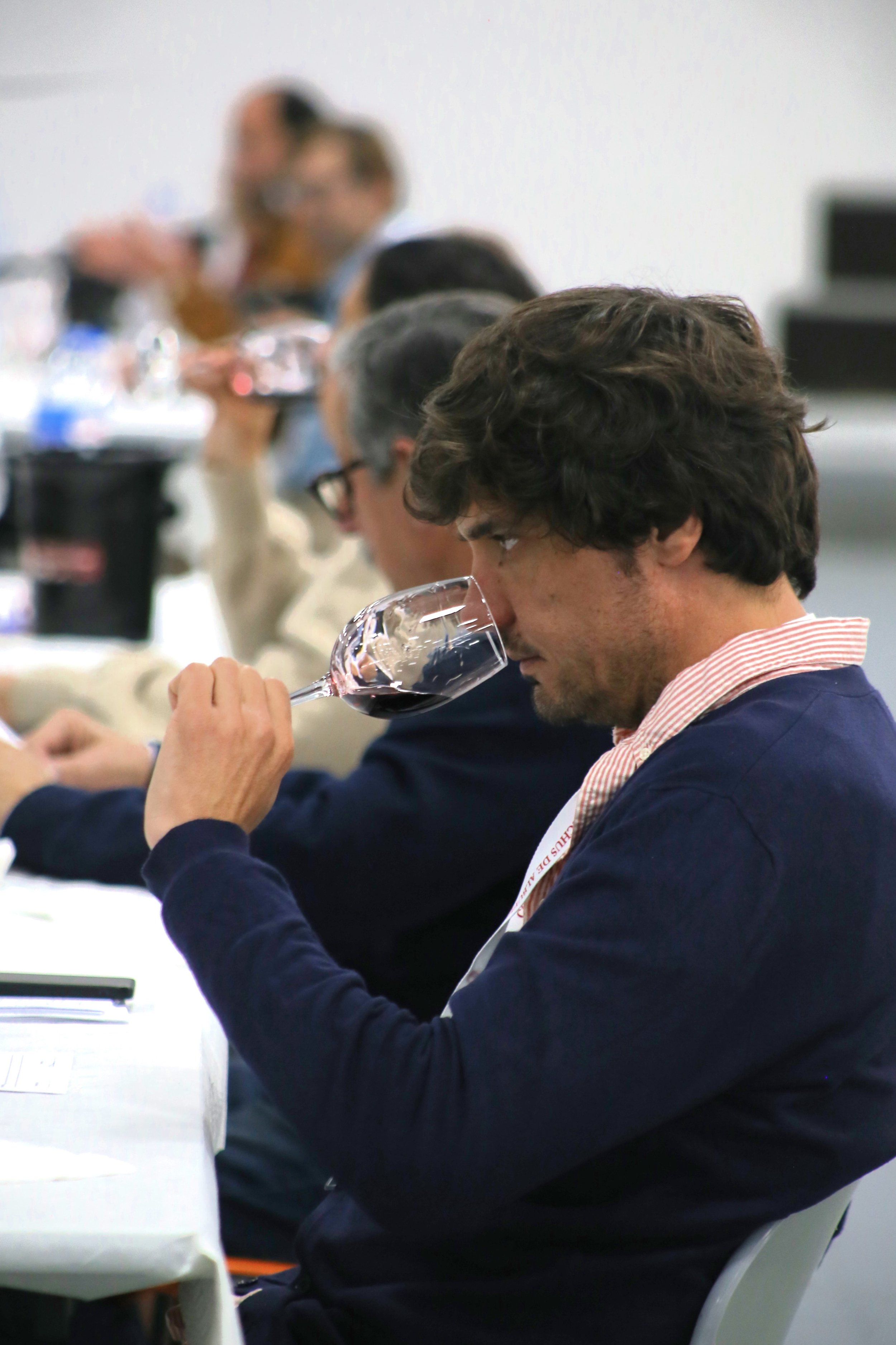 Homem com cabelo castanho escuro e barba de nível médio, usando camisa de botão e suéter azul escuro, cheirando taça de vinho tinto em uma mesa de uma degustação de vinhos, com outras pessoas ao fundo também degustando vinho.