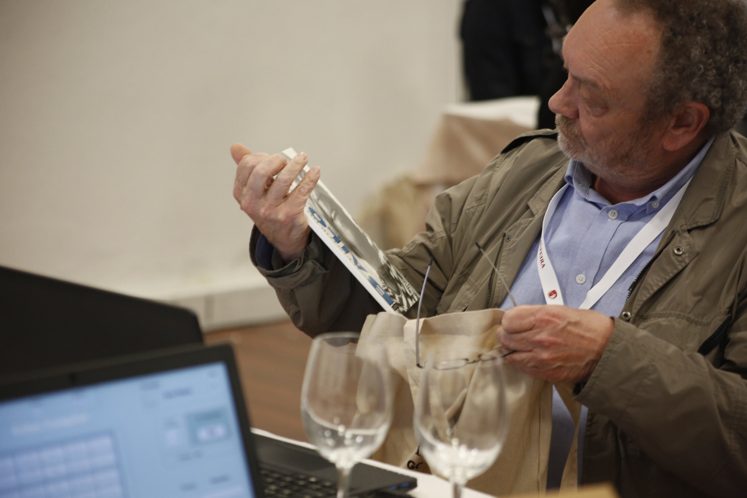 Homem de cabelo cacheado e barba, usando jaqueta e camisa azul, segurando uma revista e um saco de papel na mão, sentado em uma mesa com duas taças de vinho vazias e um laptop.