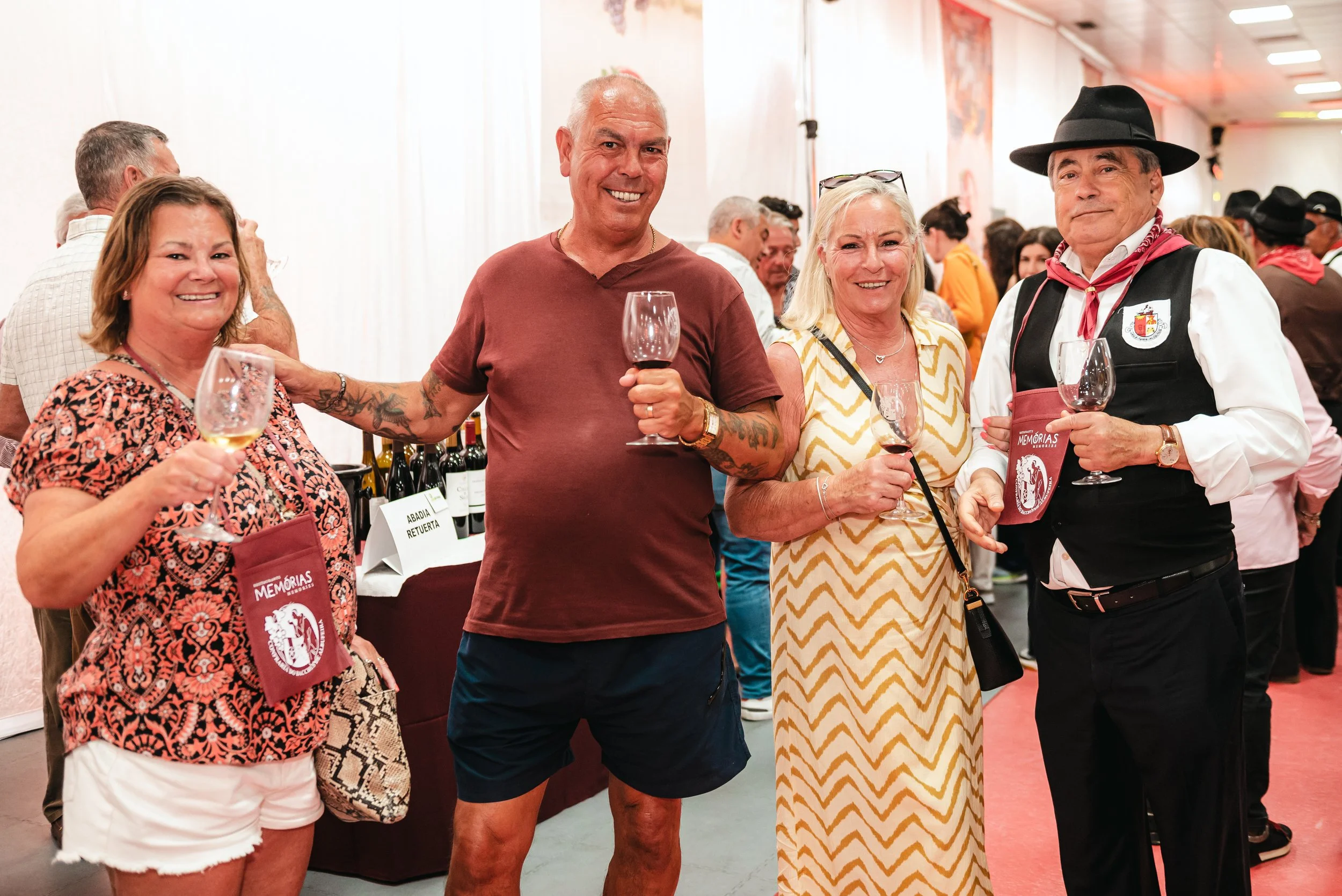 Quatro pessoas posando com taças de vinho em evento social, em um ambiente interno com várias pessoas ao fundo.