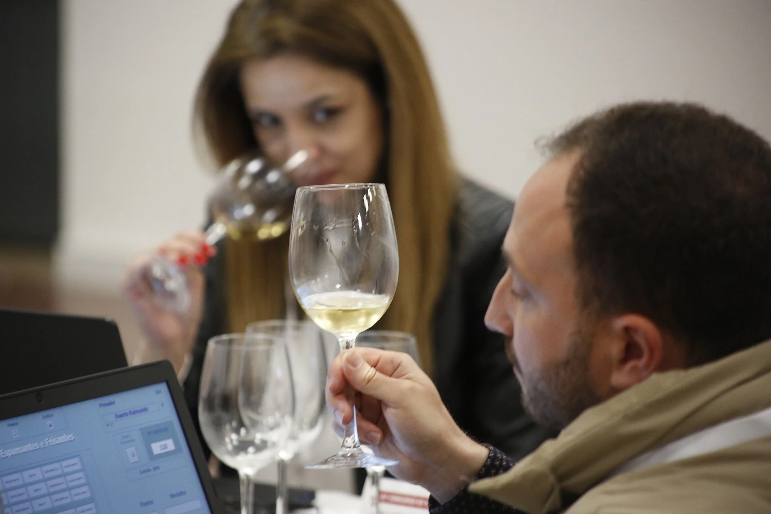 Homem segurando taça de vinho branco, mulher ao fundo com taça de vinho, em uma mesa com várias taças de vinho e laptop.
