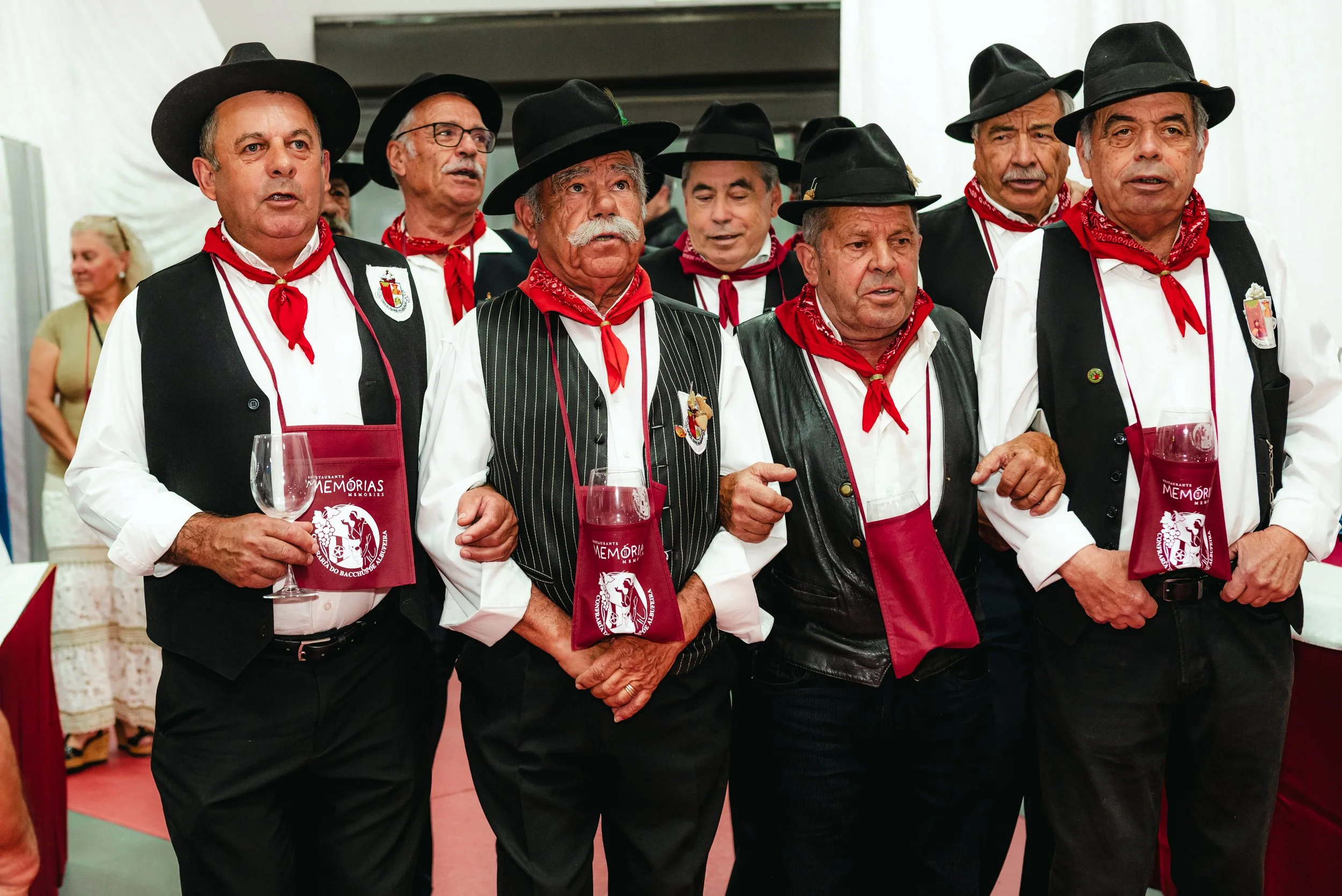 Grupo de homens idoso vestindo roupas tradicionais, chapéus pretos, coletes, lenços vermelhos no pescoço, segurando taças, participando de evento cultural.