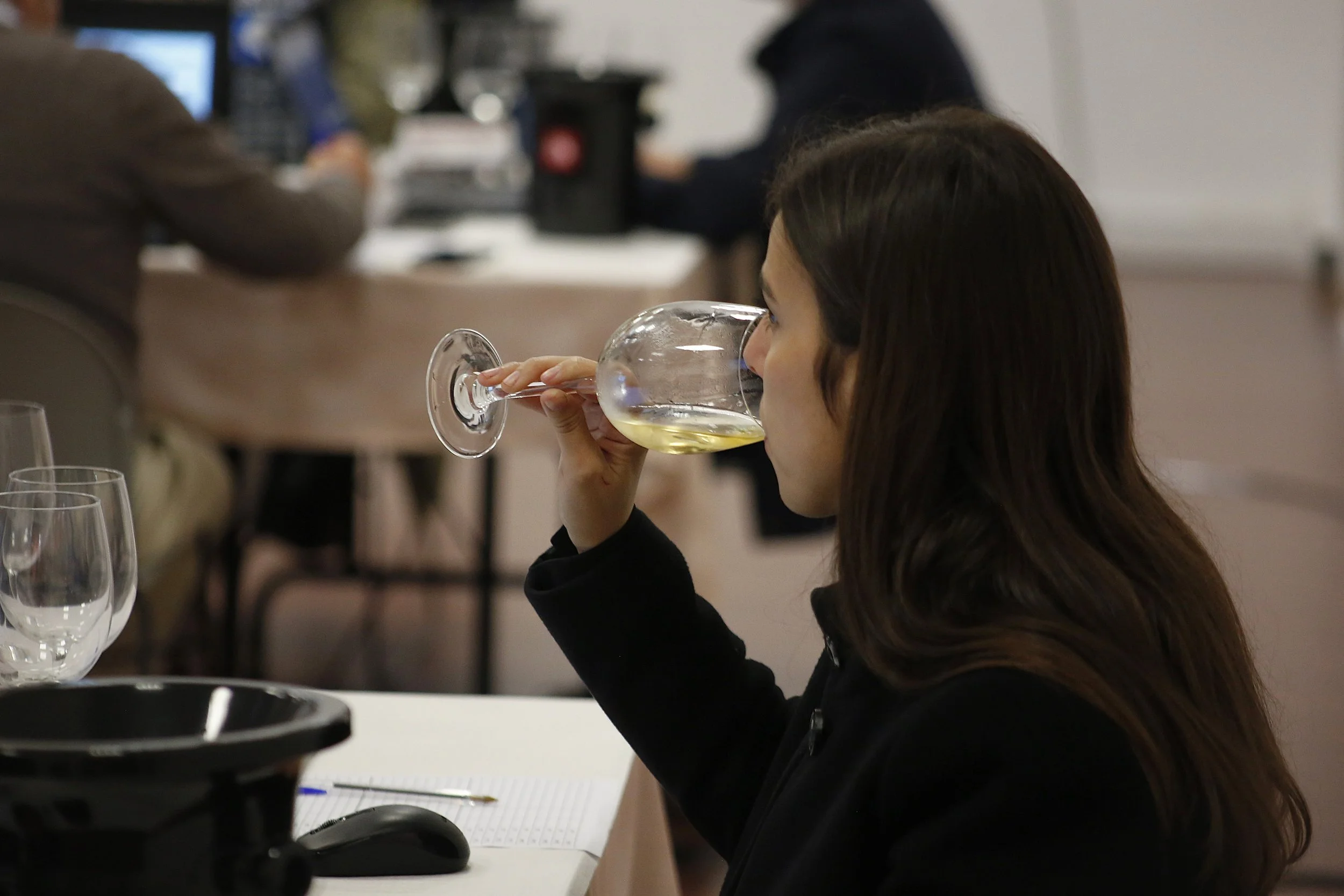 Mulher de cabelos castanhos escuros bebendo vinho branco em taça em uma reunião ou evento.