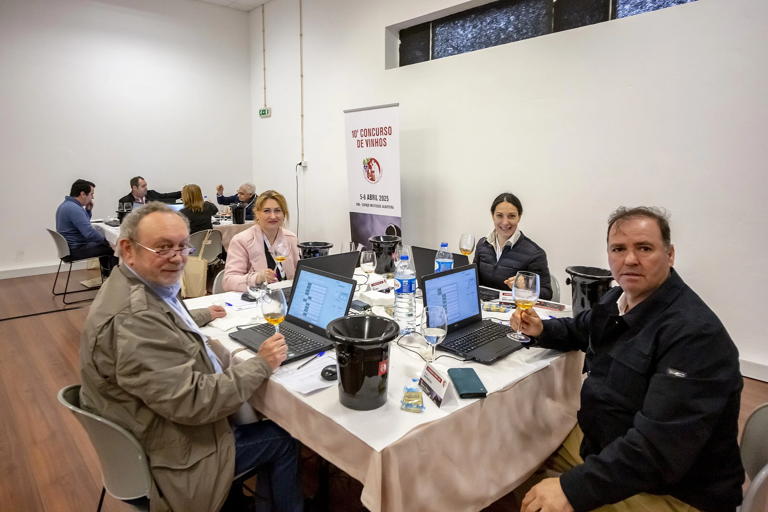 Reunião de pessoas sentadas ao redor de uma mesa em um evento de concurso de vinhos, com laptops, taças de vinho e garrafas de água, em uma sala interna