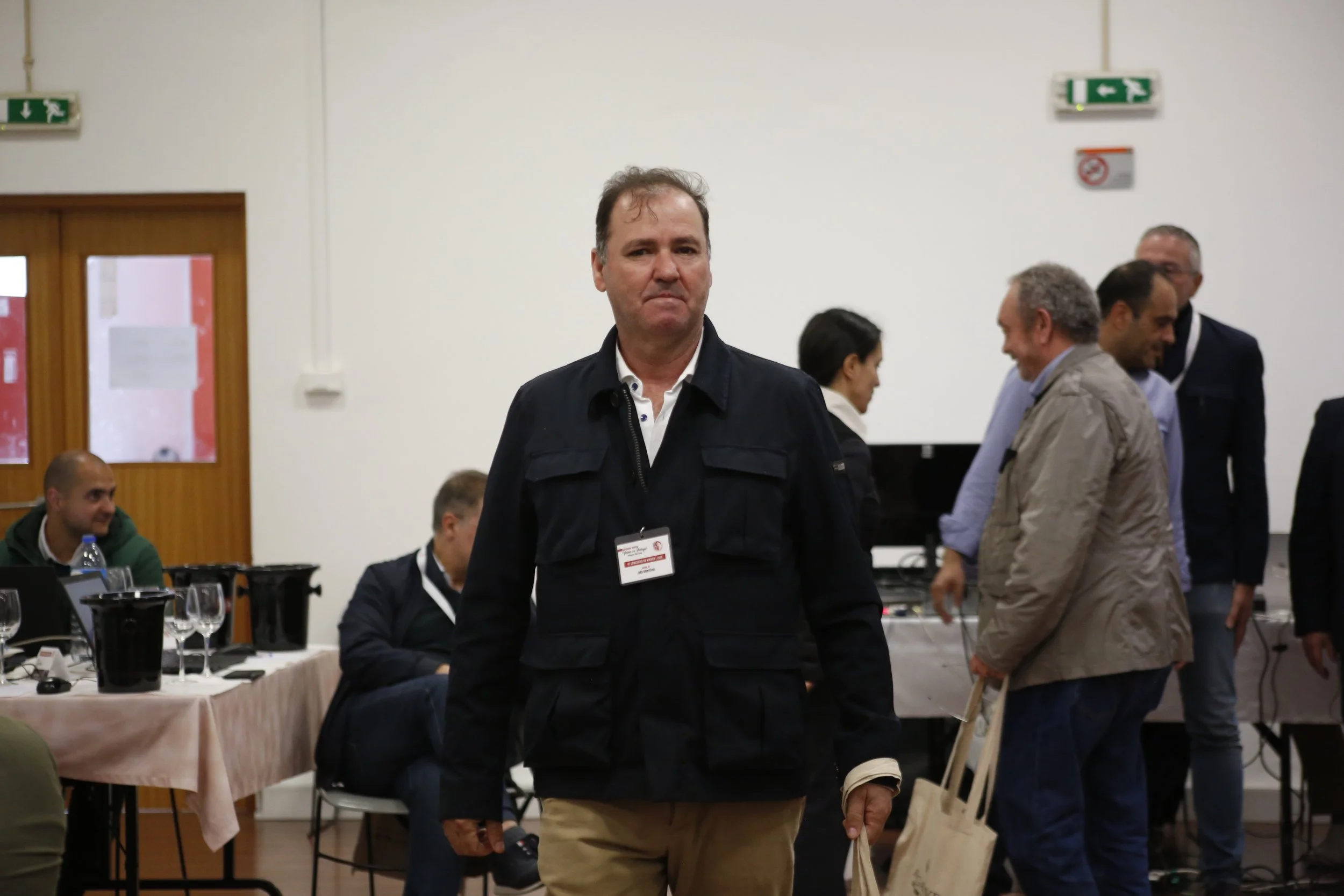 Homem de terno casual em uma conferência ou reunião, segurando uma sacola de papel, com várias pessoas ao fundo conversando e mesas com copos e garrafas de água.