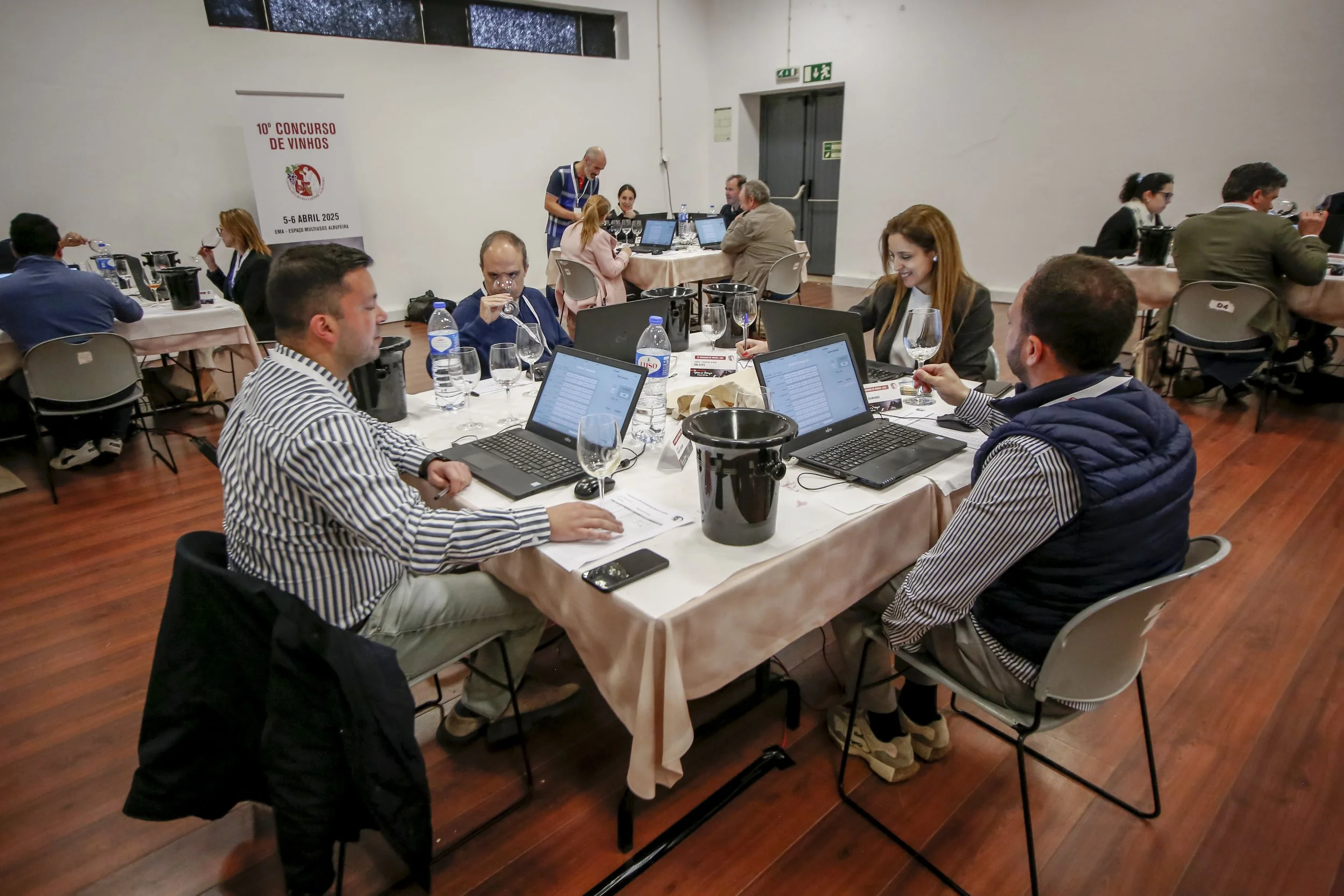 Participantes reunidos em uma sala de conferências, com laptops, copos de vinho e garrafas de água em uma mesa, durante o 10º Concurso de Vinhos, realizado em abril de 2025.
