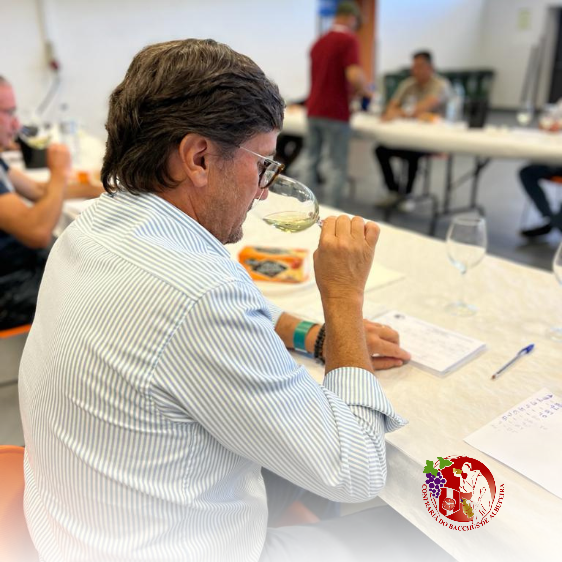 Homem degustando vinho em uma mesa, com várias pessoas ao fundo, em um ambiente de confraria de vinho