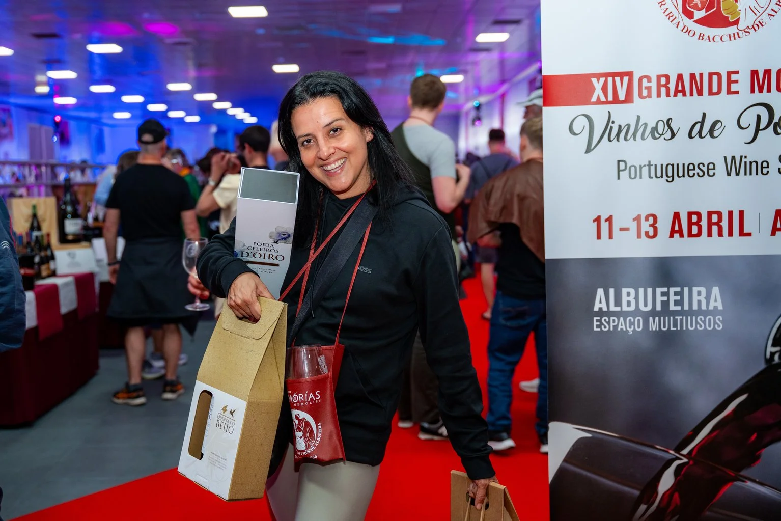 Mulher sorridente segurando várias caixas de vinho em um evento de degustação, ao lado de um painel com informações sobre o evento